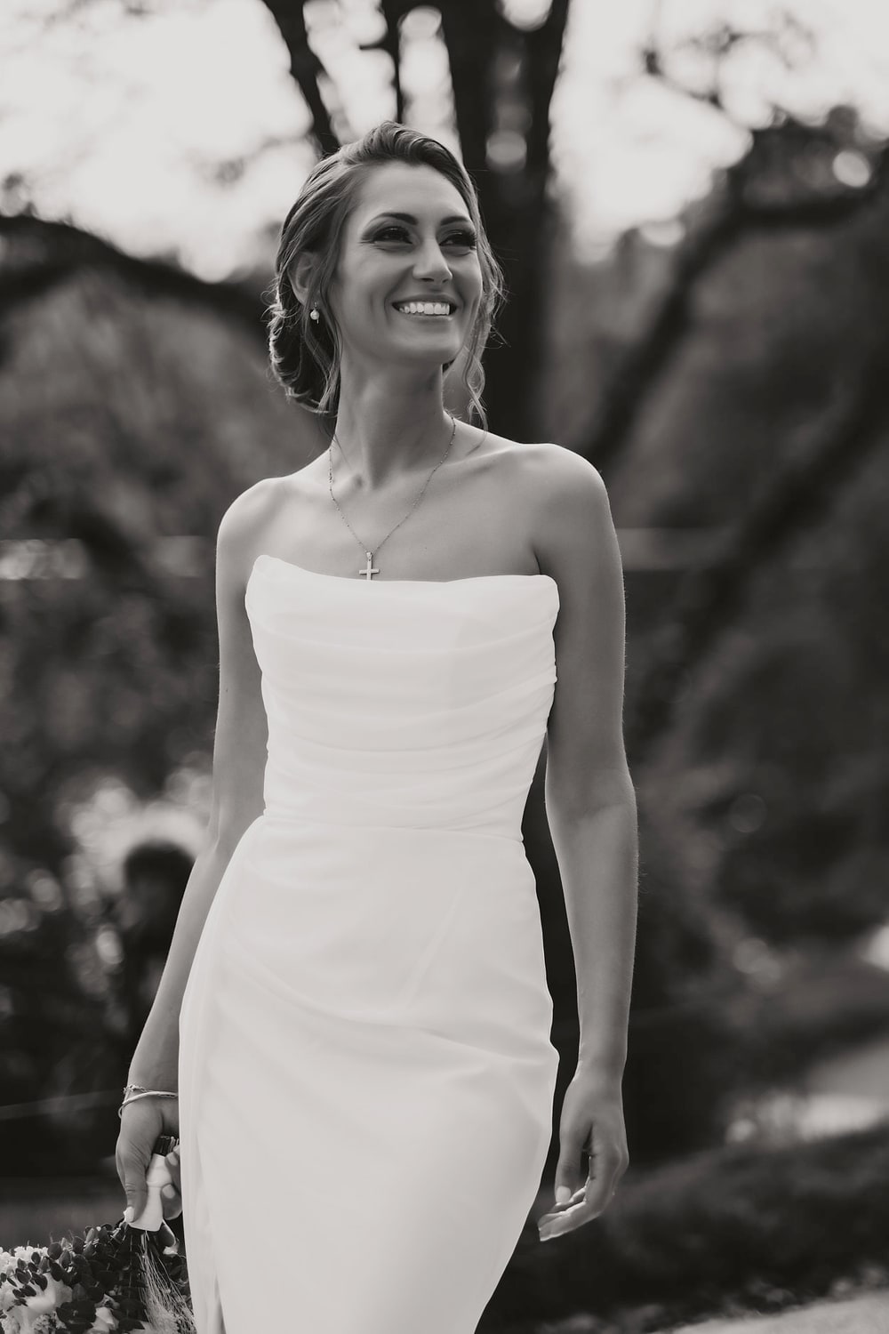 A bride in a plain satin dress standing among trees, looking off to one side
