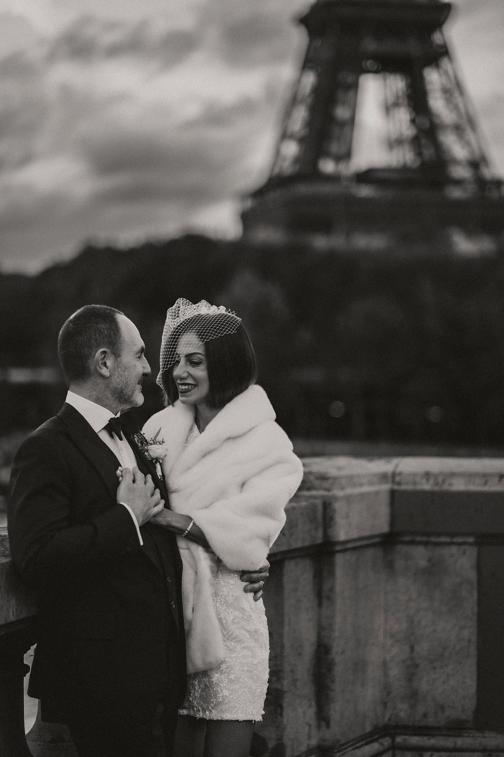 A bride in a white coat held close by her husband on a cold terrace above the Seine