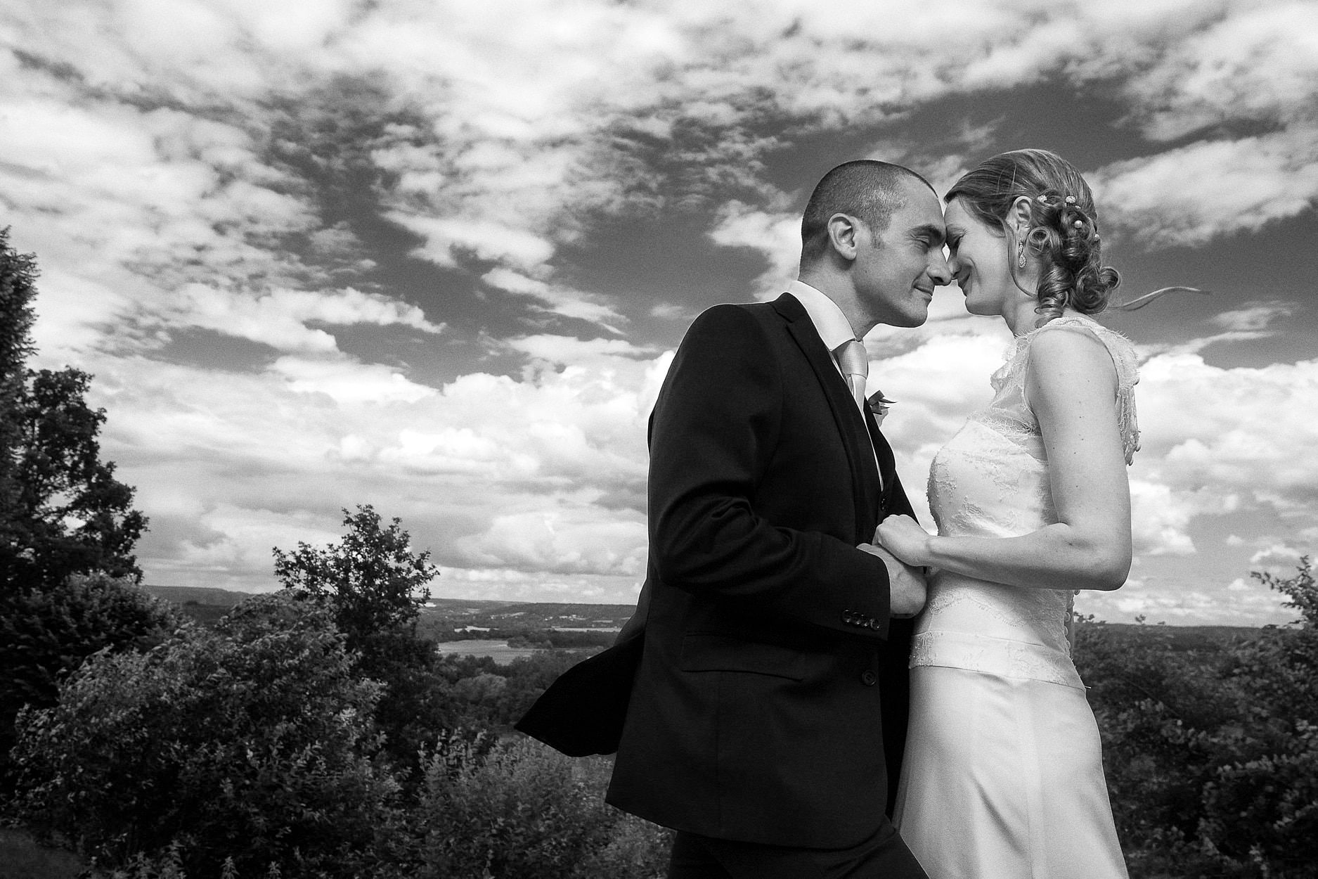 A bride and groom forehead to forehead in an open field under a wide cloudy sky