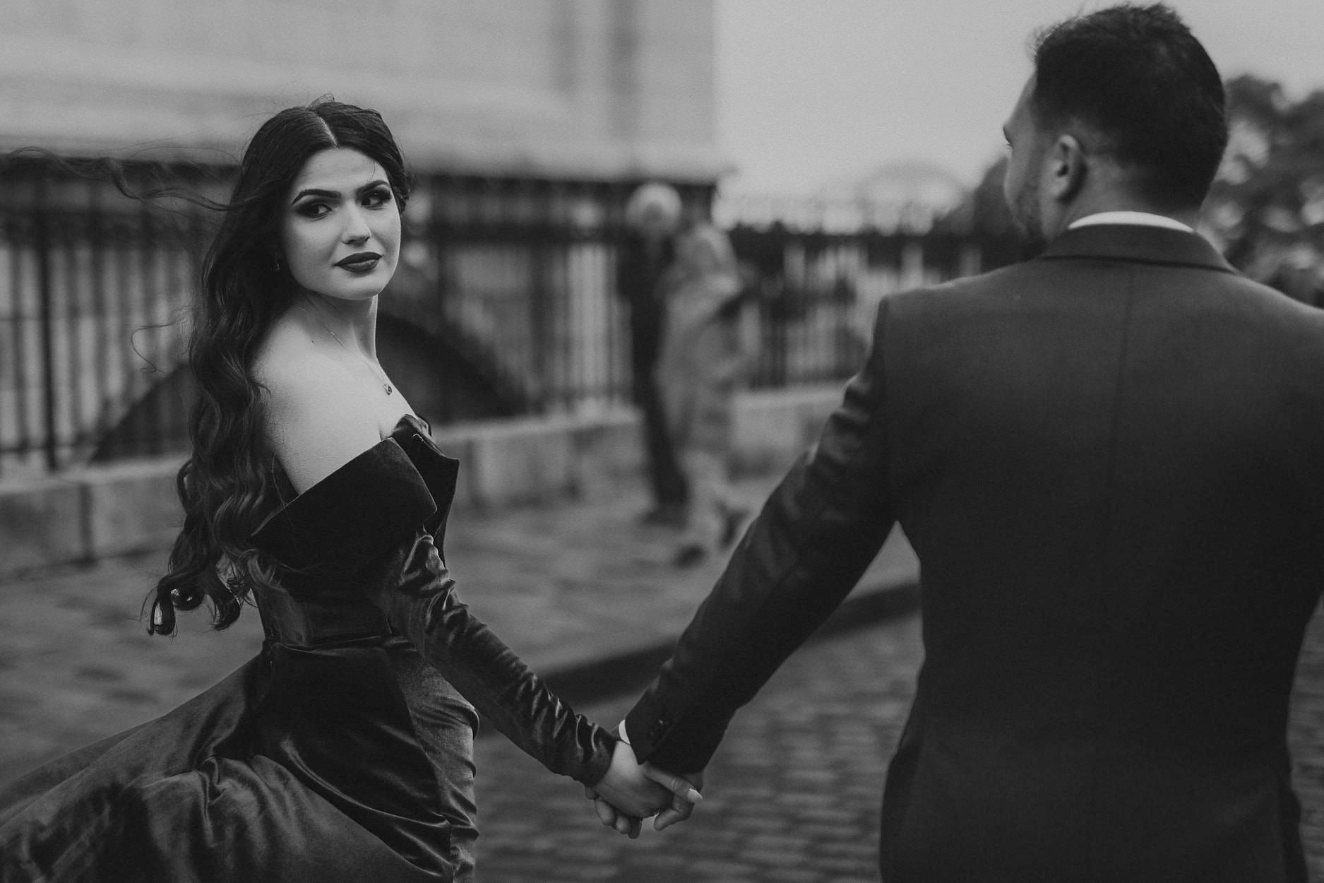 A woman in a black velvet gown looks back over her shoulder as she is led by the hand along a cobbled street in Montmartre
