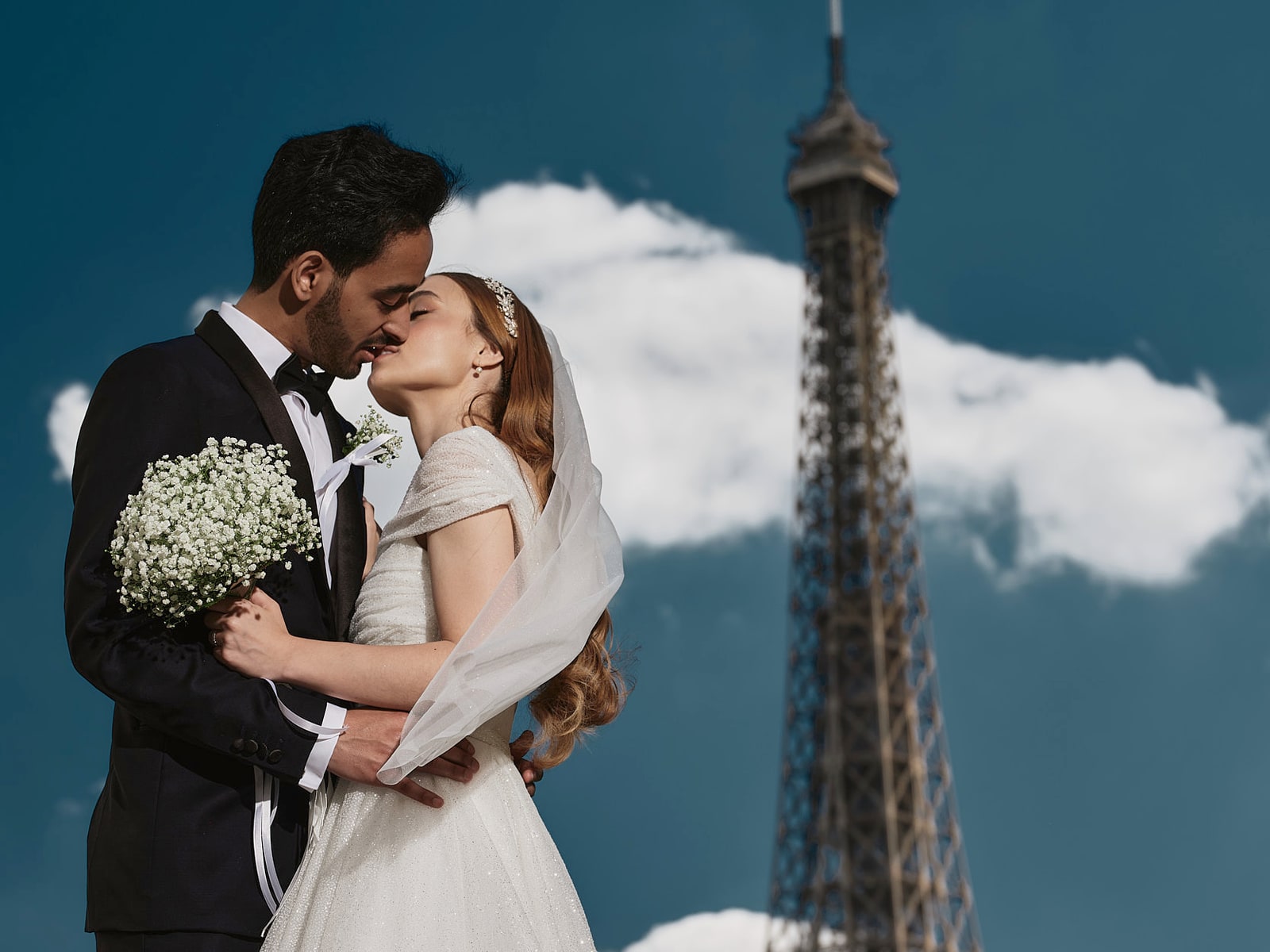A bride and groom kissing, her veil catching the light and a bouquet of gypsophila between them, with the Eiffel Tower rising behind against a deep blue sky