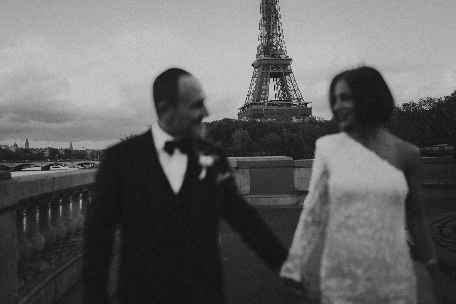 A bride and groom holding hands on a terrace above the Seine, the Eiffel Tower rising between them