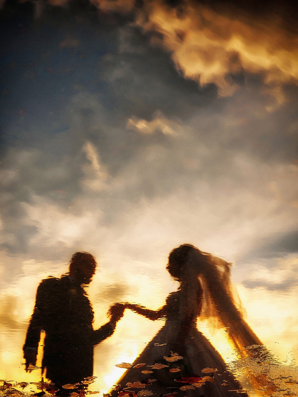 A bride and groom reflected in a rain puddle at sunset, reaching for each other’s hands against a burning orange sky