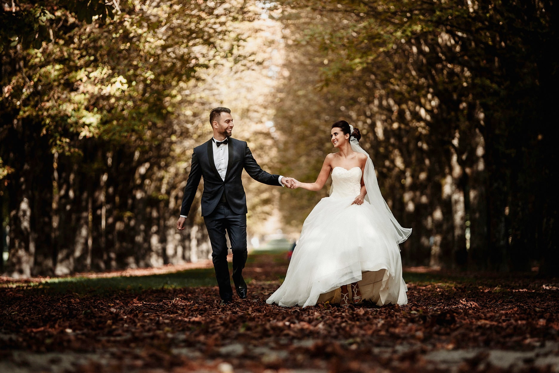 A groom leading his bride down an avenue of trees in low autumn light