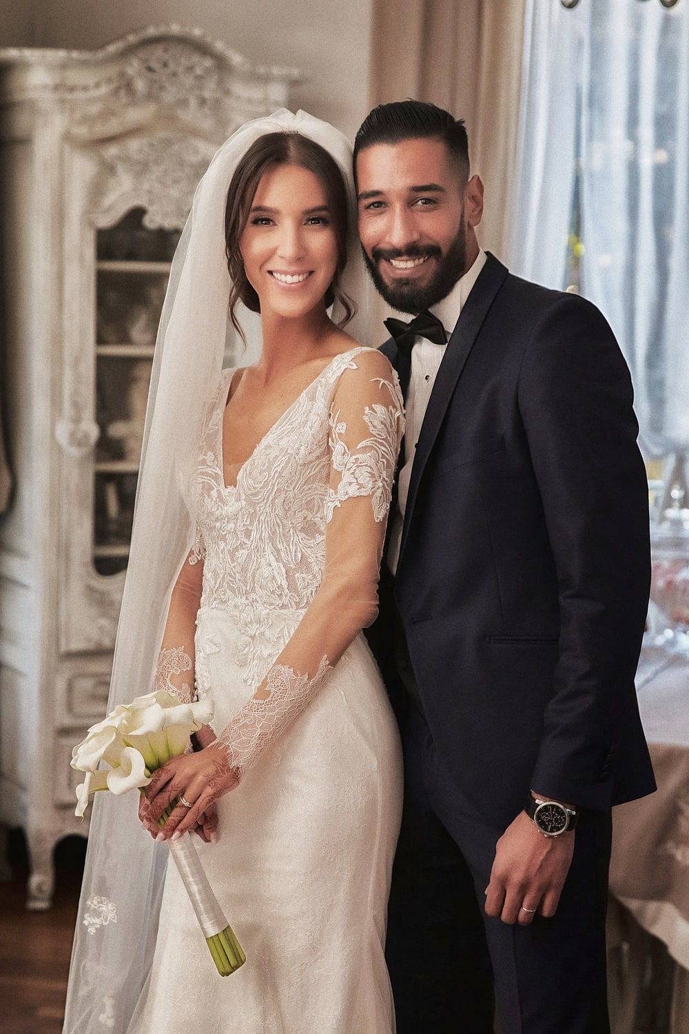 A bride and groom standing side by side in front of a bright window, both looking straight at the camera