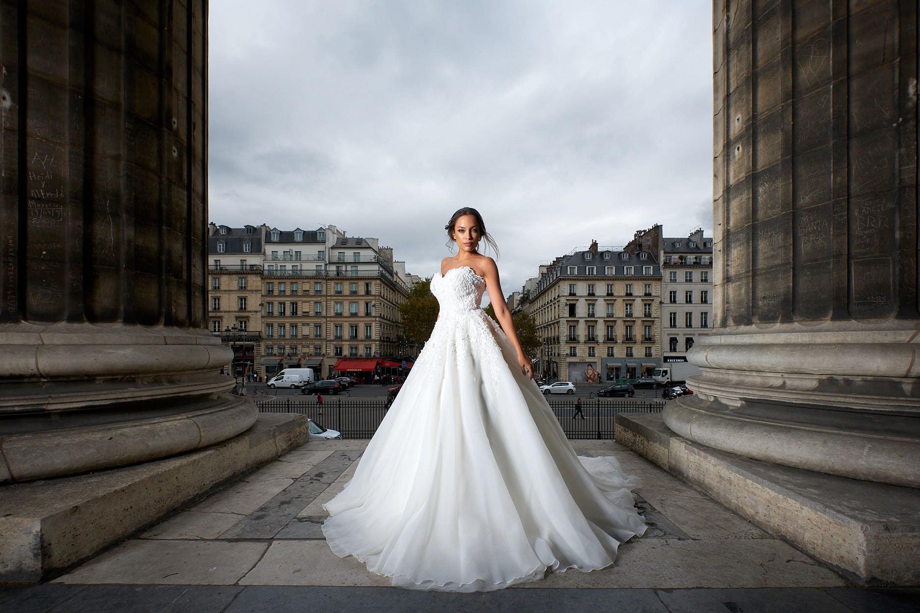 A bride standing between two enormous stone columns, the Paris street stretching away beyond her