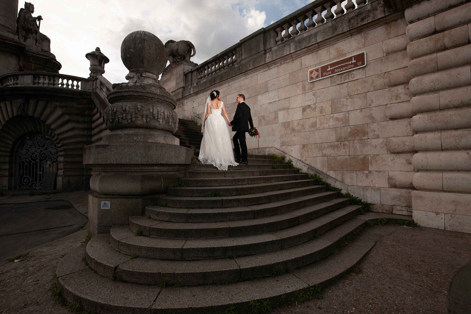 A bride and groom climbing the curved stone steps up to the Pont Alexandre III, hand in hand, the balustrade and its carved lion above them
