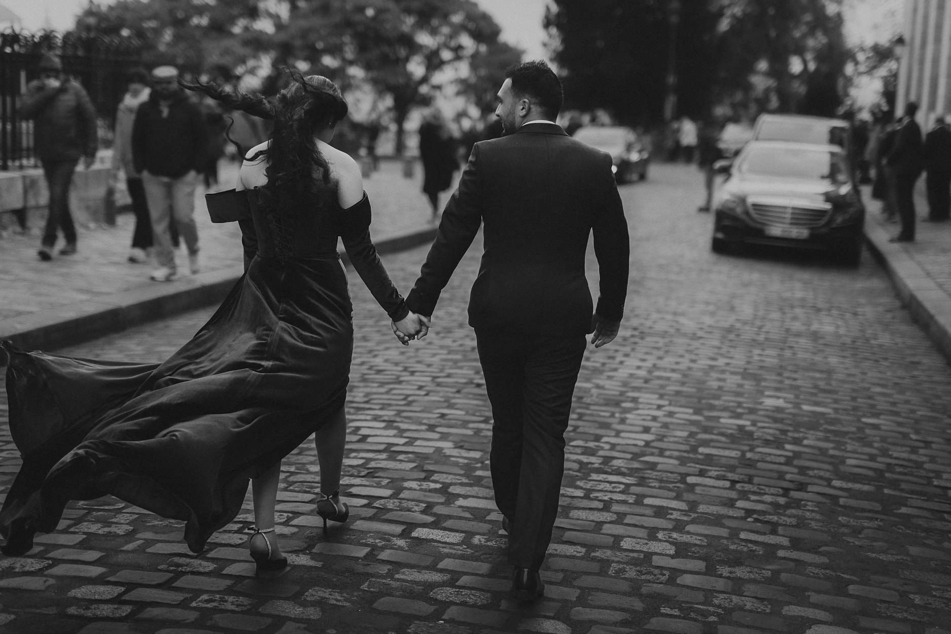 A couple crossing wet cobblestones hand in hand, her long black skirt lifting behind her