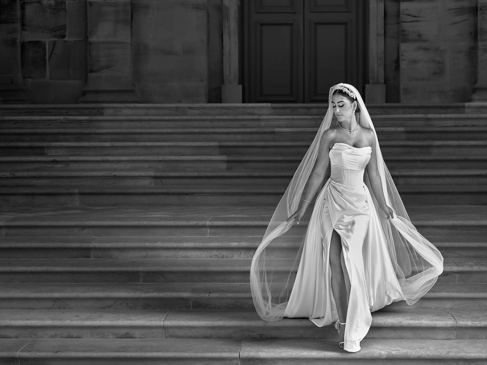 A bride walking down a wide flight of stone steps in black and white, her veil trailing far behind her