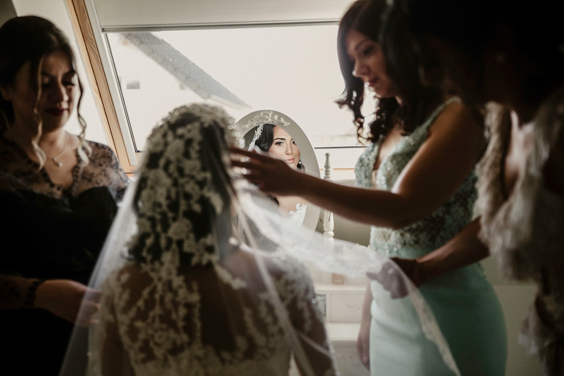 Two women lifting a bride’s veil into place at a bright window