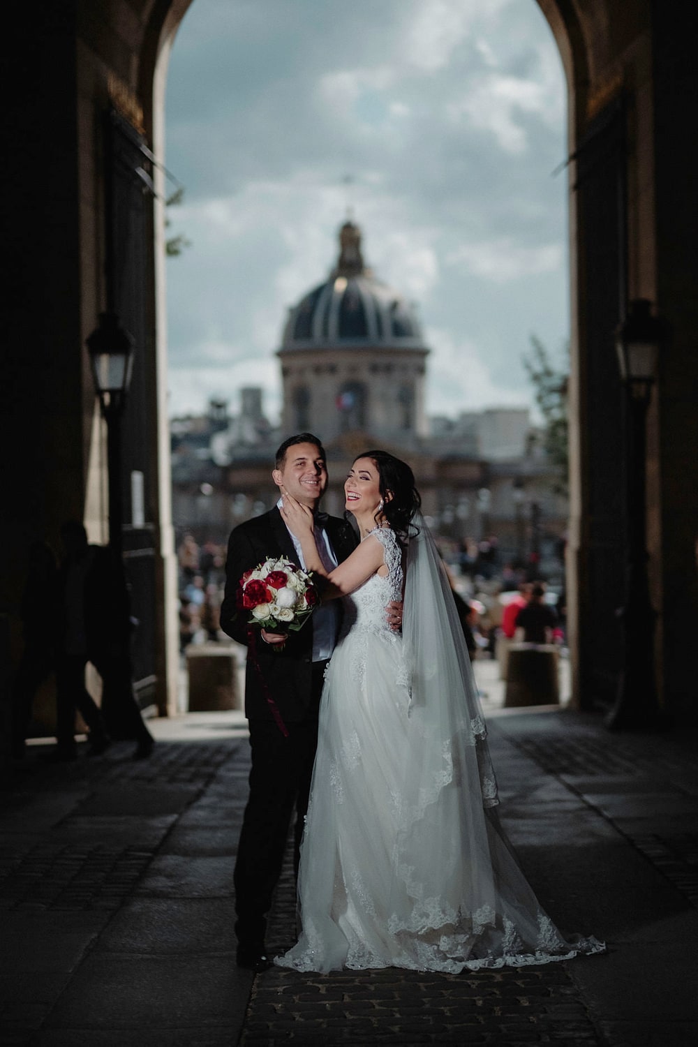 A couple standing in a stone archway that frames the dome of the Institut de France across the river