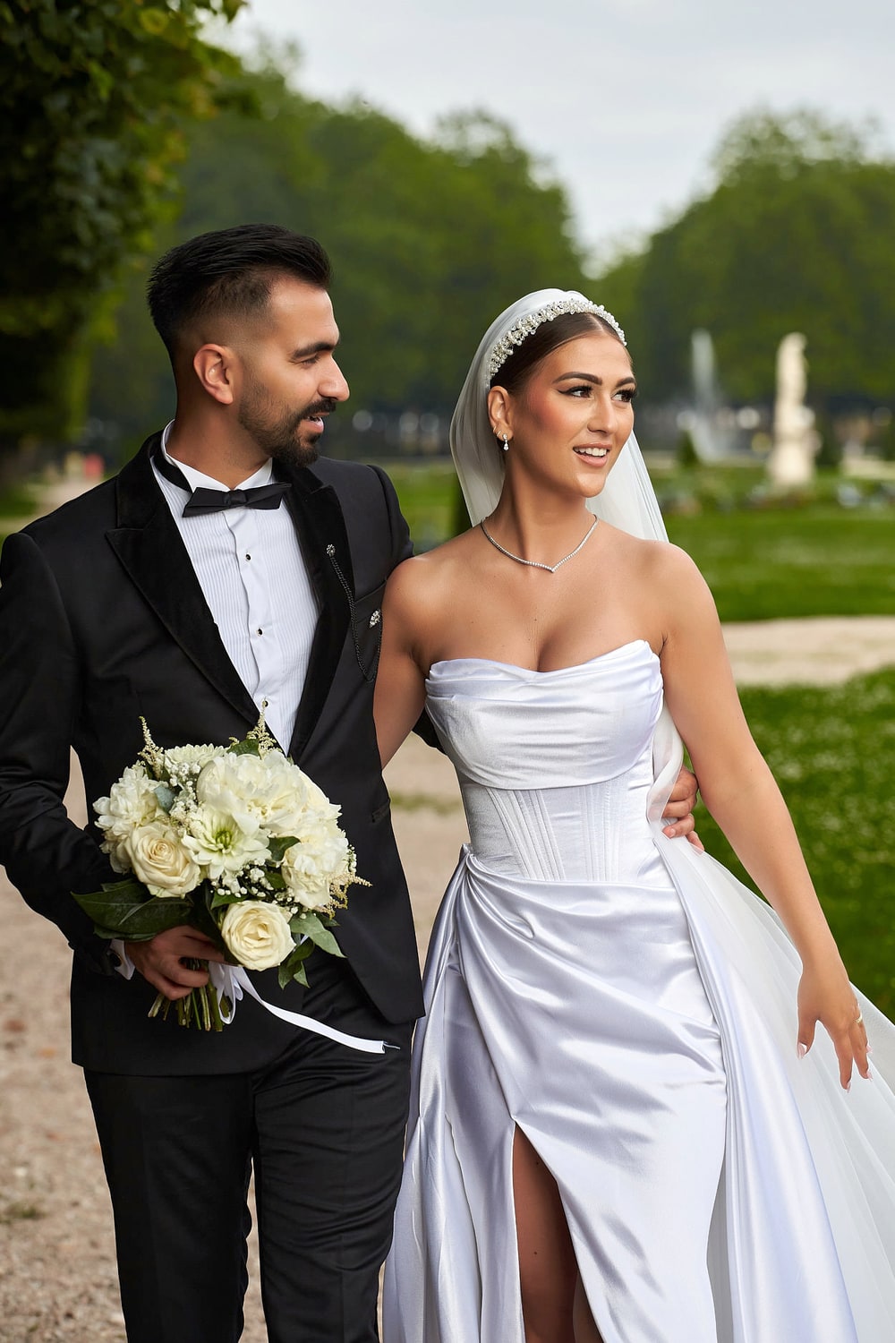 A couple standing together in formal gardens, the groom holding a bouquet of white roses