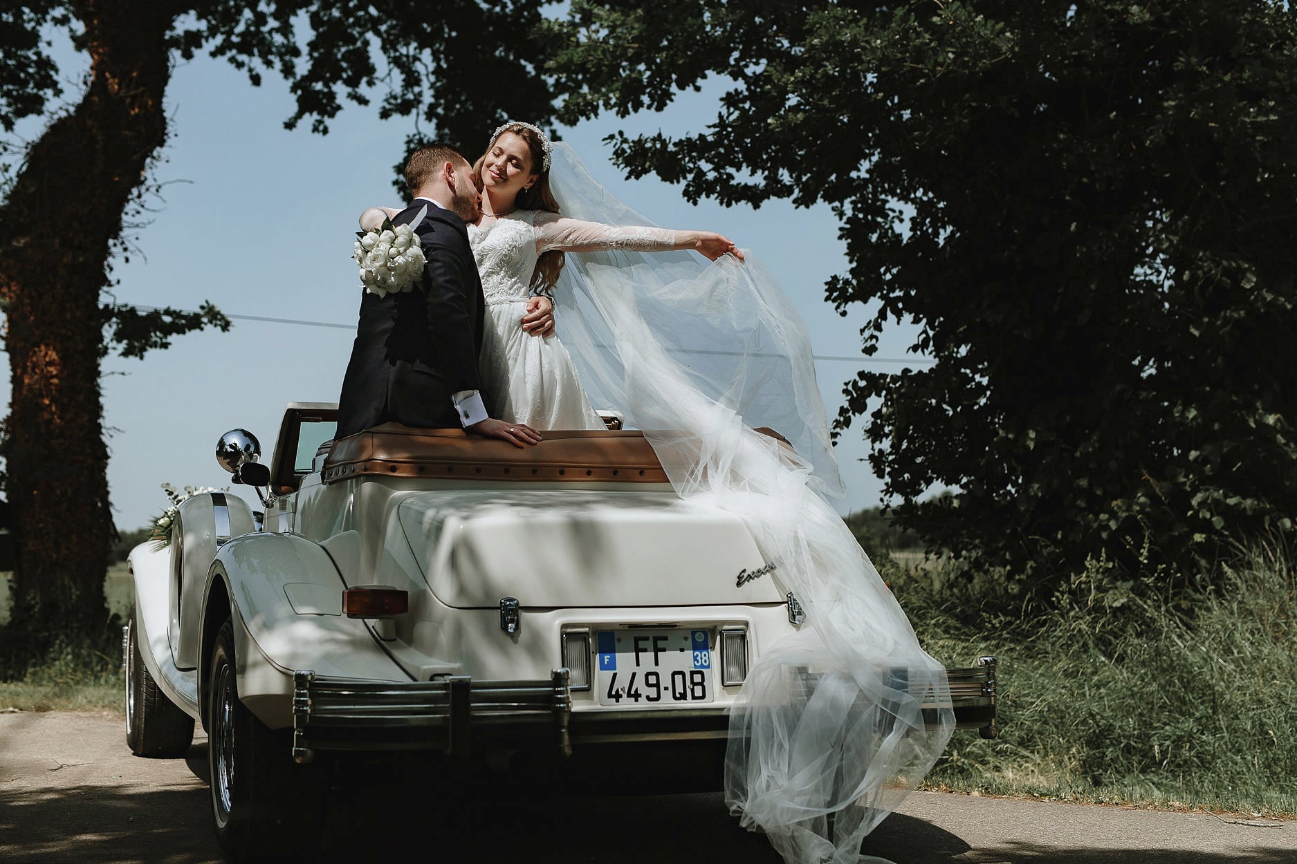 A bride and groom standing up in an open vintage car, her veil streaming behind them