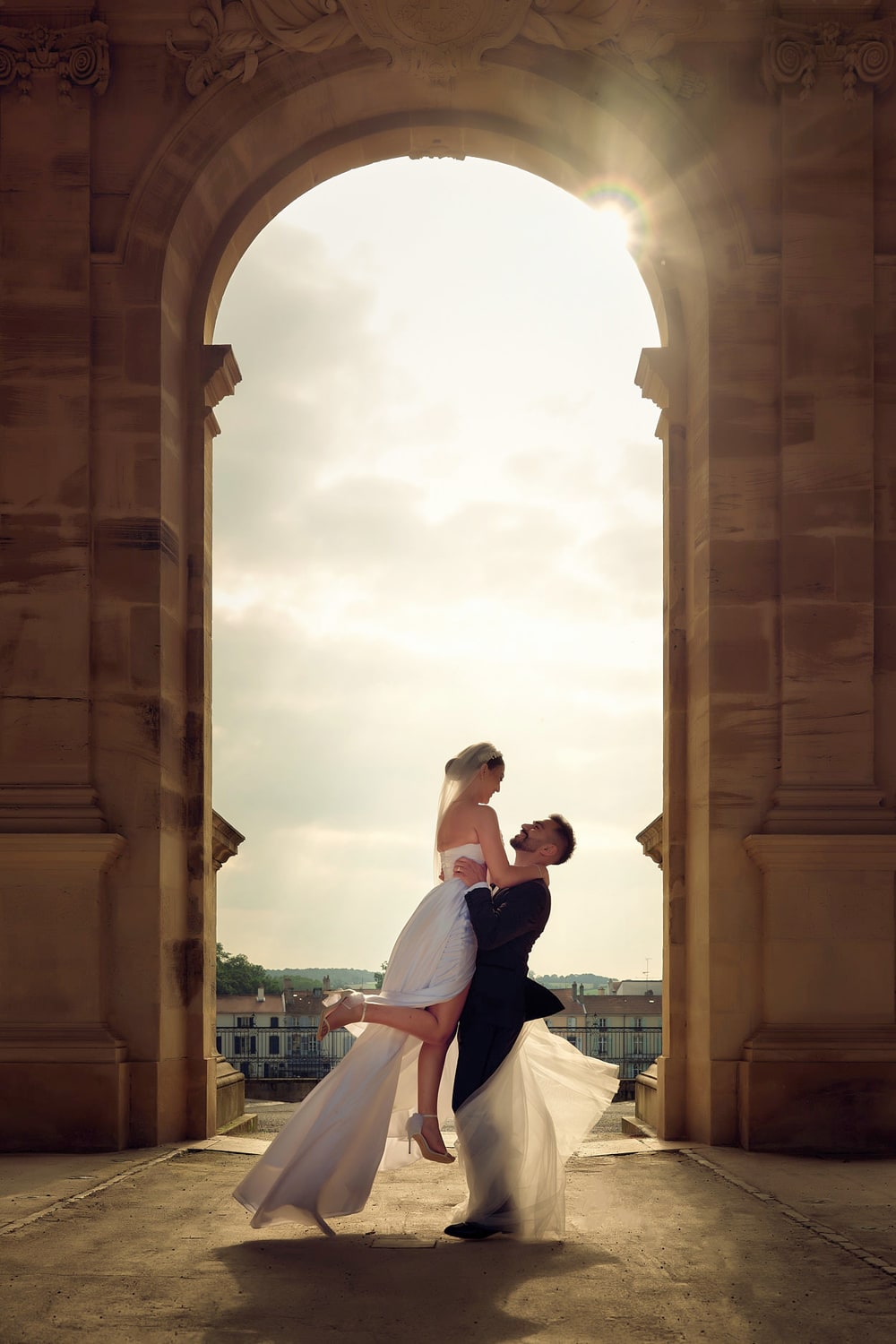 A groom lifting his bride in a sunlit stone archway, the city visible beyond
