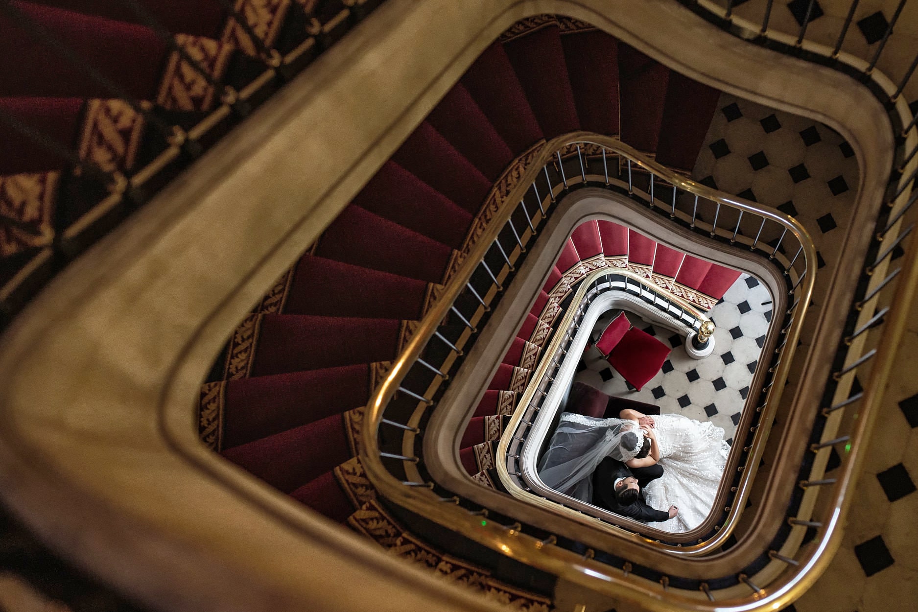 A spiral staircase photographed from the top floor, the couple tiny at the bottom of it