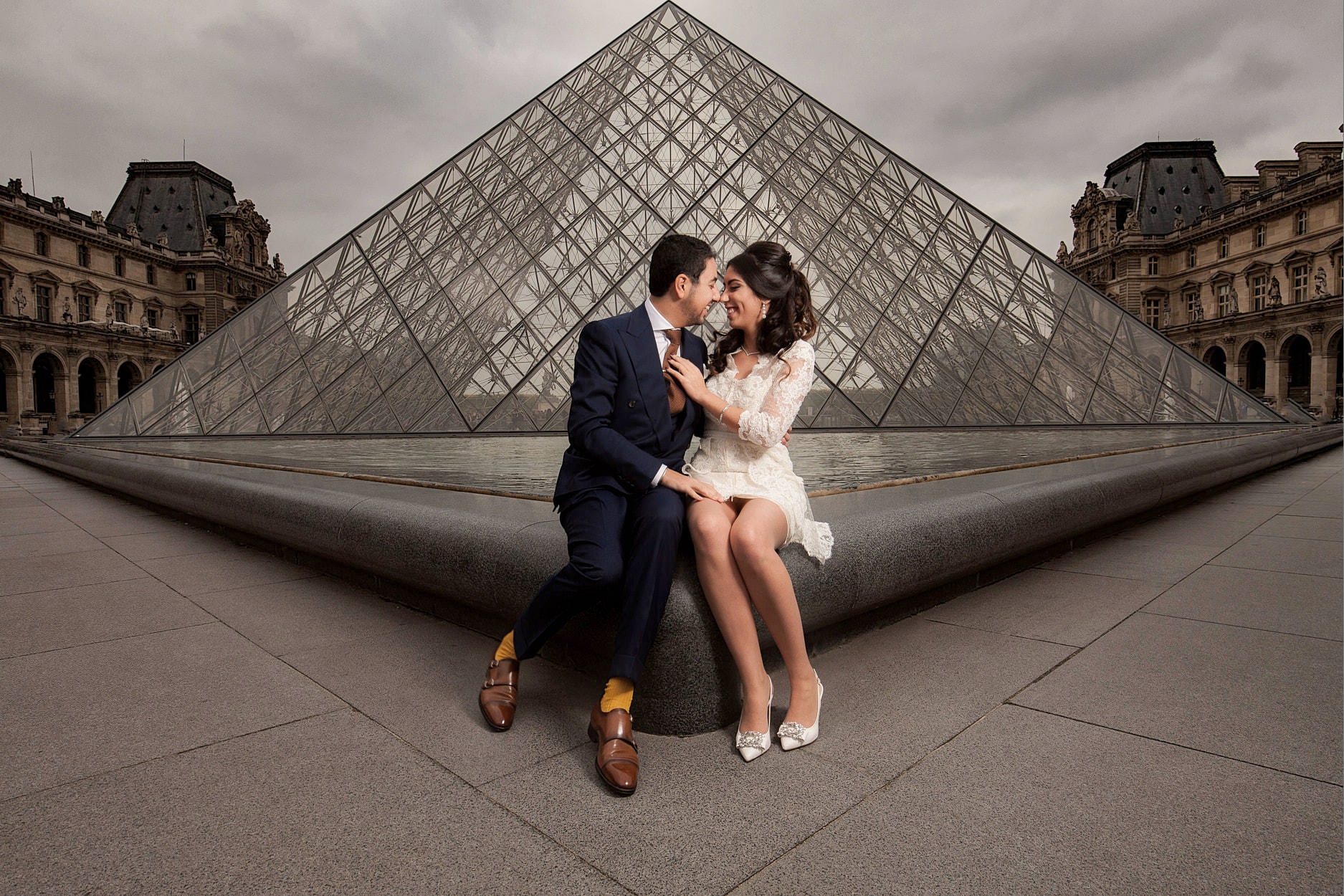 A couple sitting together on the stone of the Louvre courtyard, the pyramid behind them