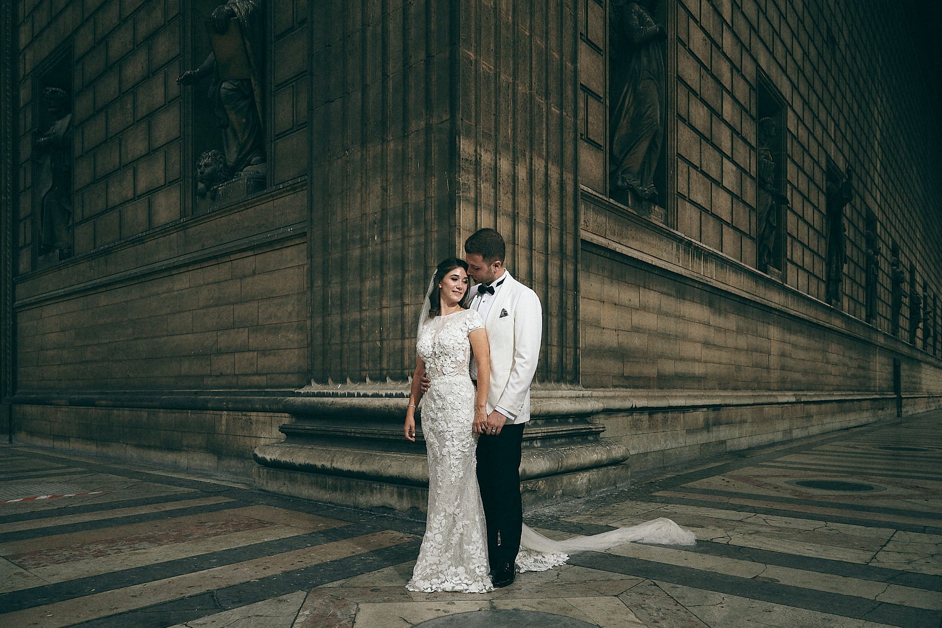 A bride and groom on the corner of a stone building, the striped paving of the courtyard running away behind them