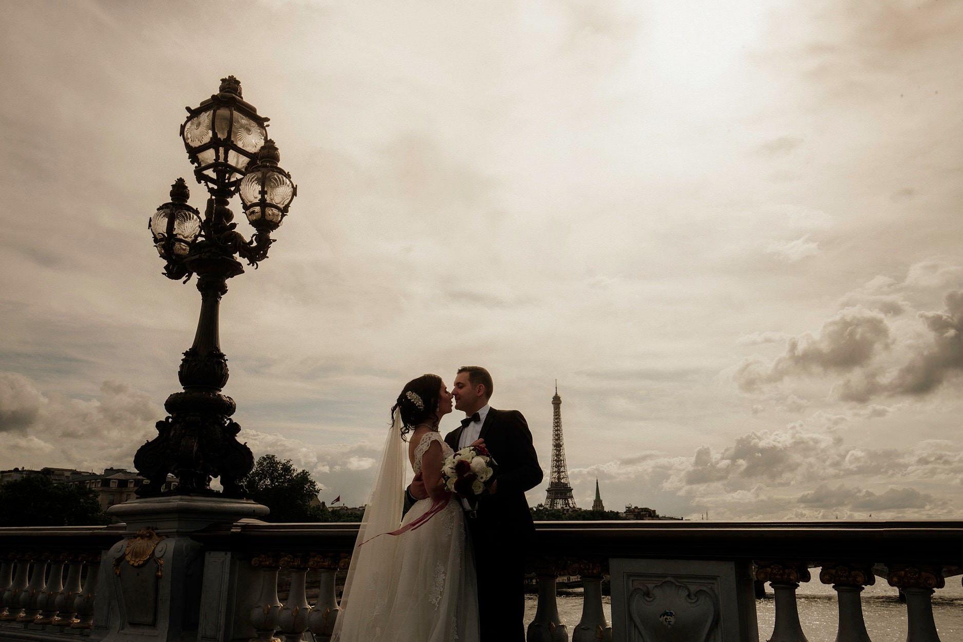 A couple kissing beneath an ornate lamp on the Pont Alexandre III, the Eiffel Tower small in the distance