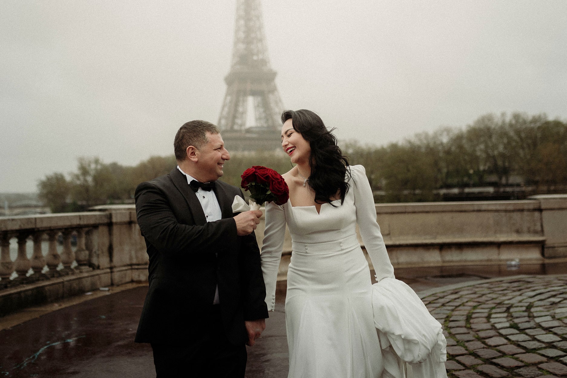 A couple facing each other on wet cobbles with red roses, the Eiffel Tower pale behind them