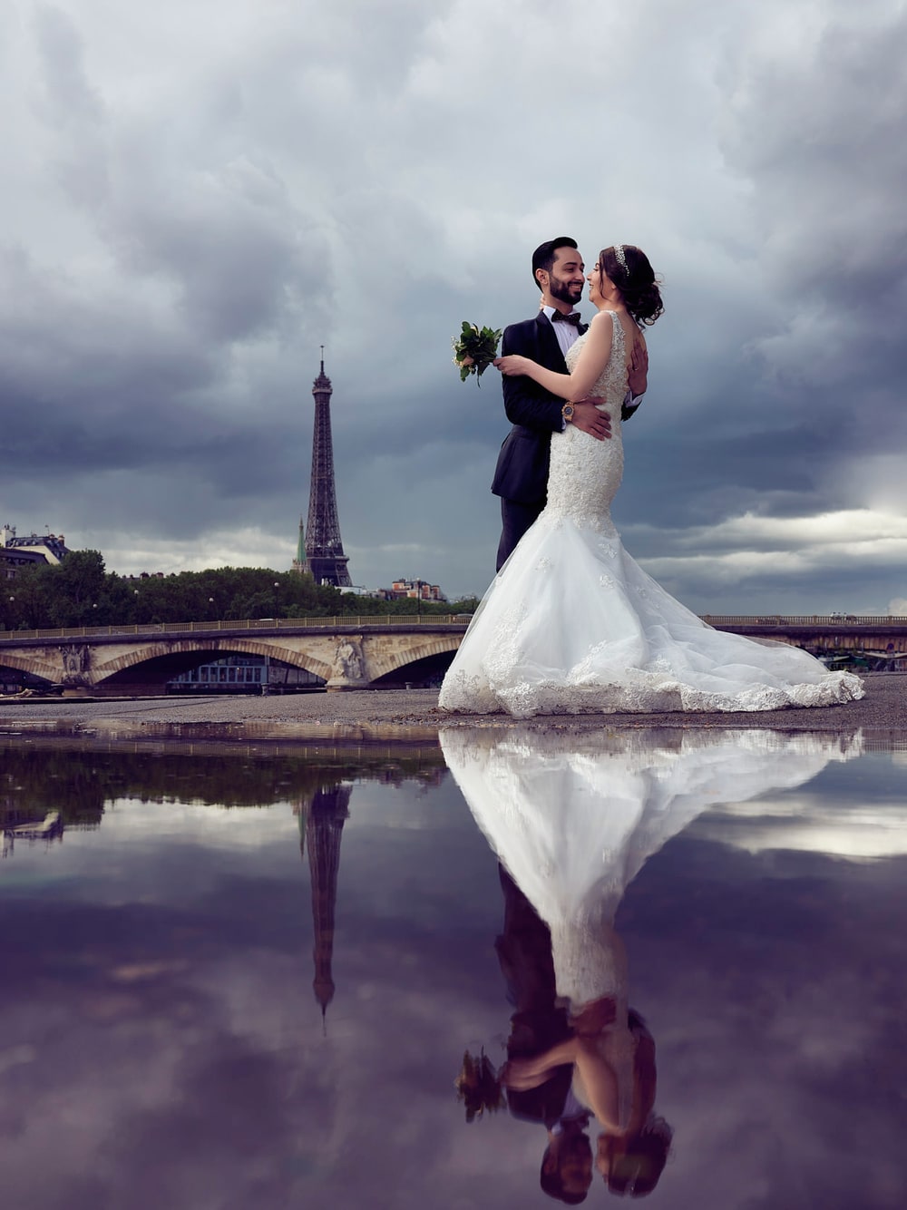 A bride and groom standing in still water, mirrored, with the Eiffel Tower behind a bank of violet cloud