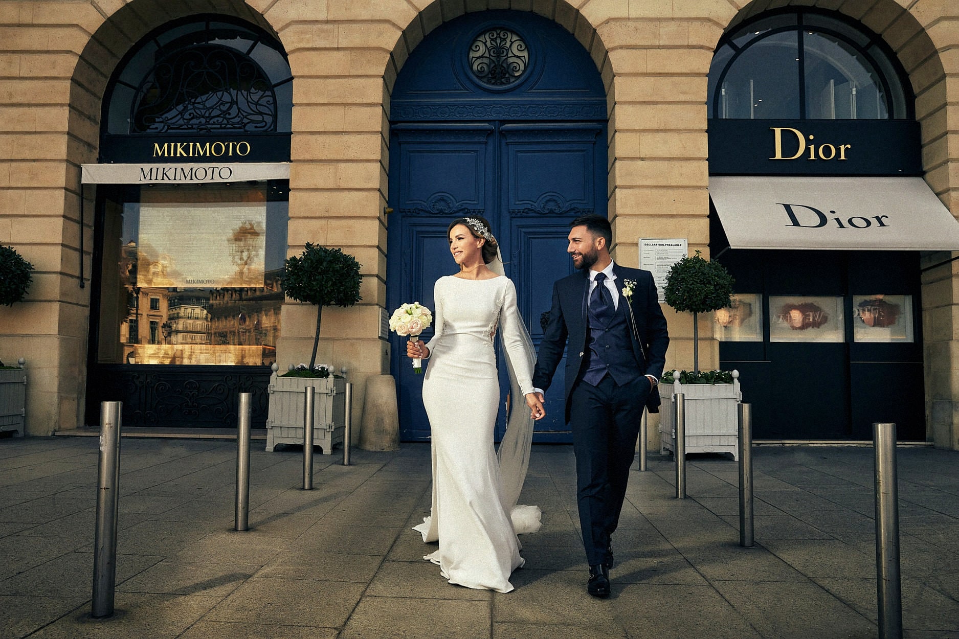 A bride and groom walking past the Dior and Mikimoto windows on a Paris shopping street