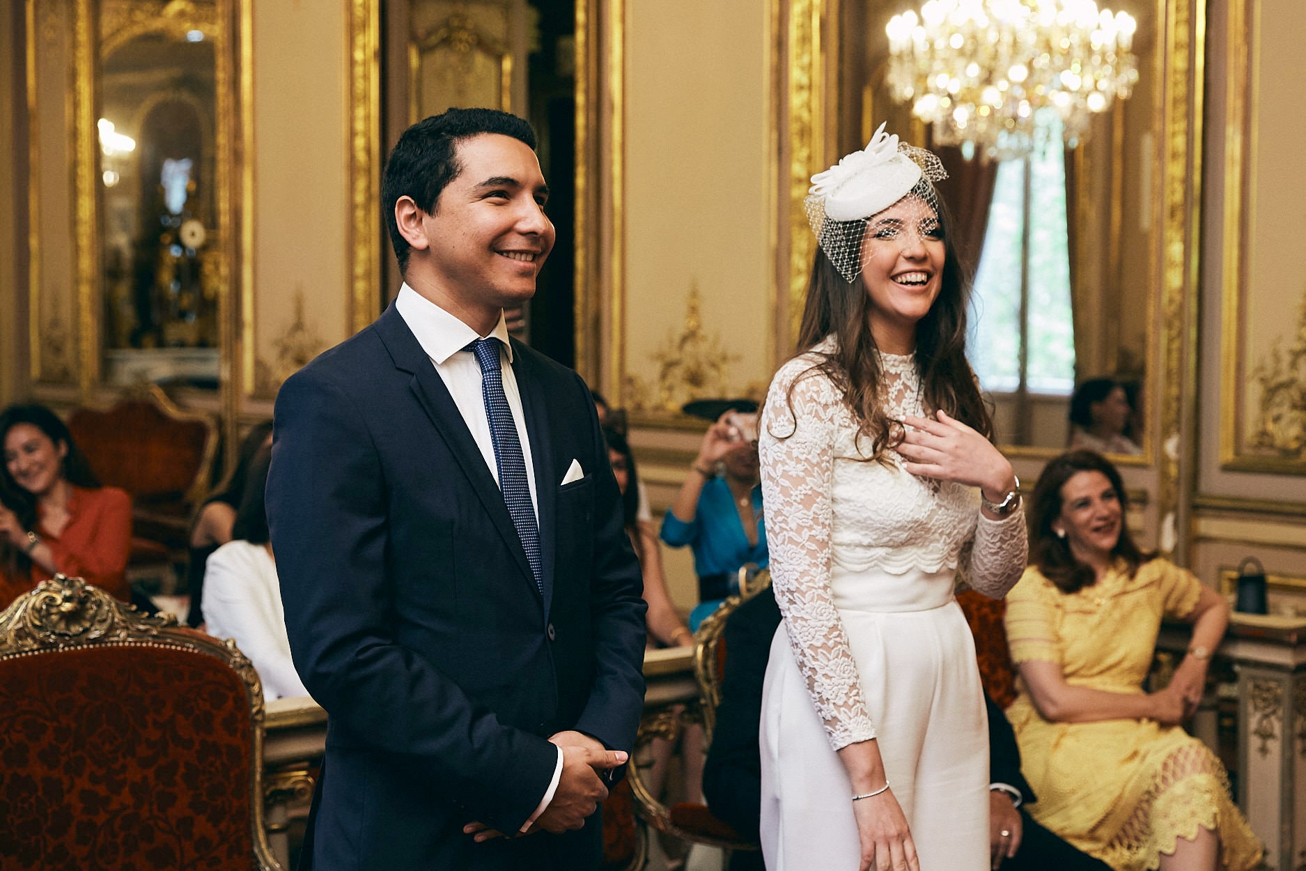 A couple standing together in the gilded ceremony room of a Paris town hall, guests seated behind them