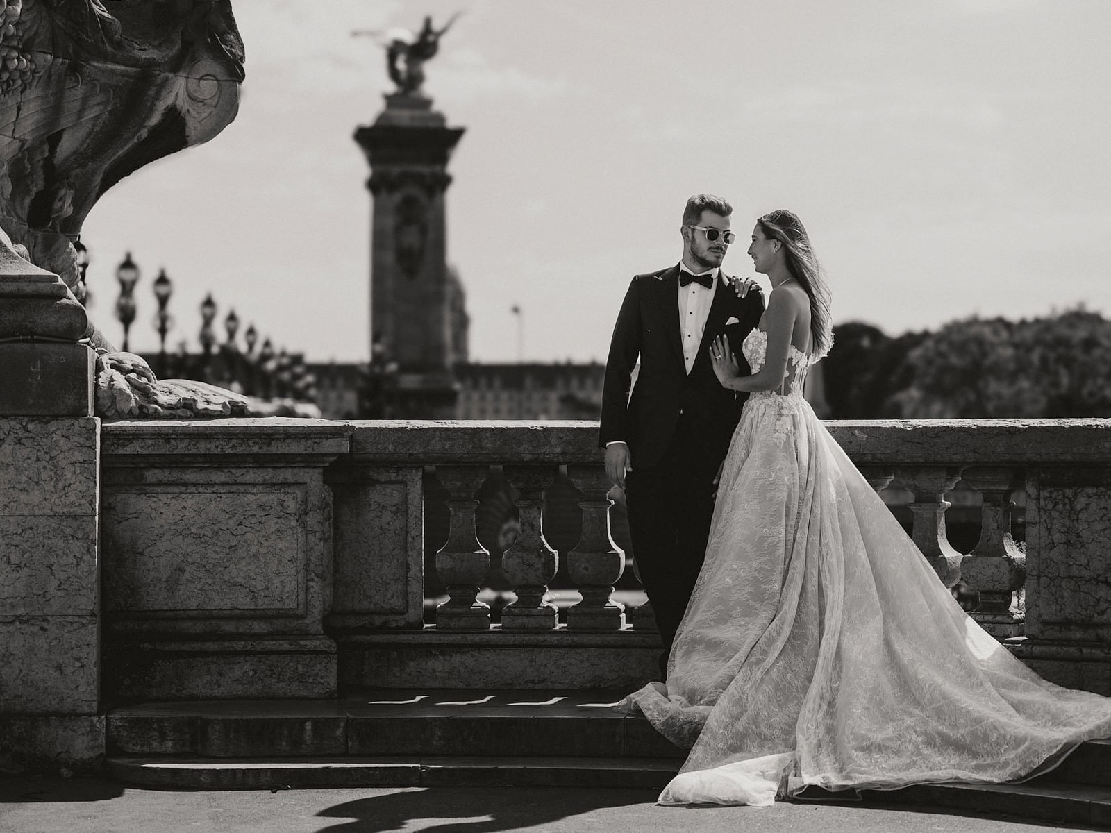 A bride and groom against the stone balustrade of the Pont Alexandre III, her train spread across the pavement