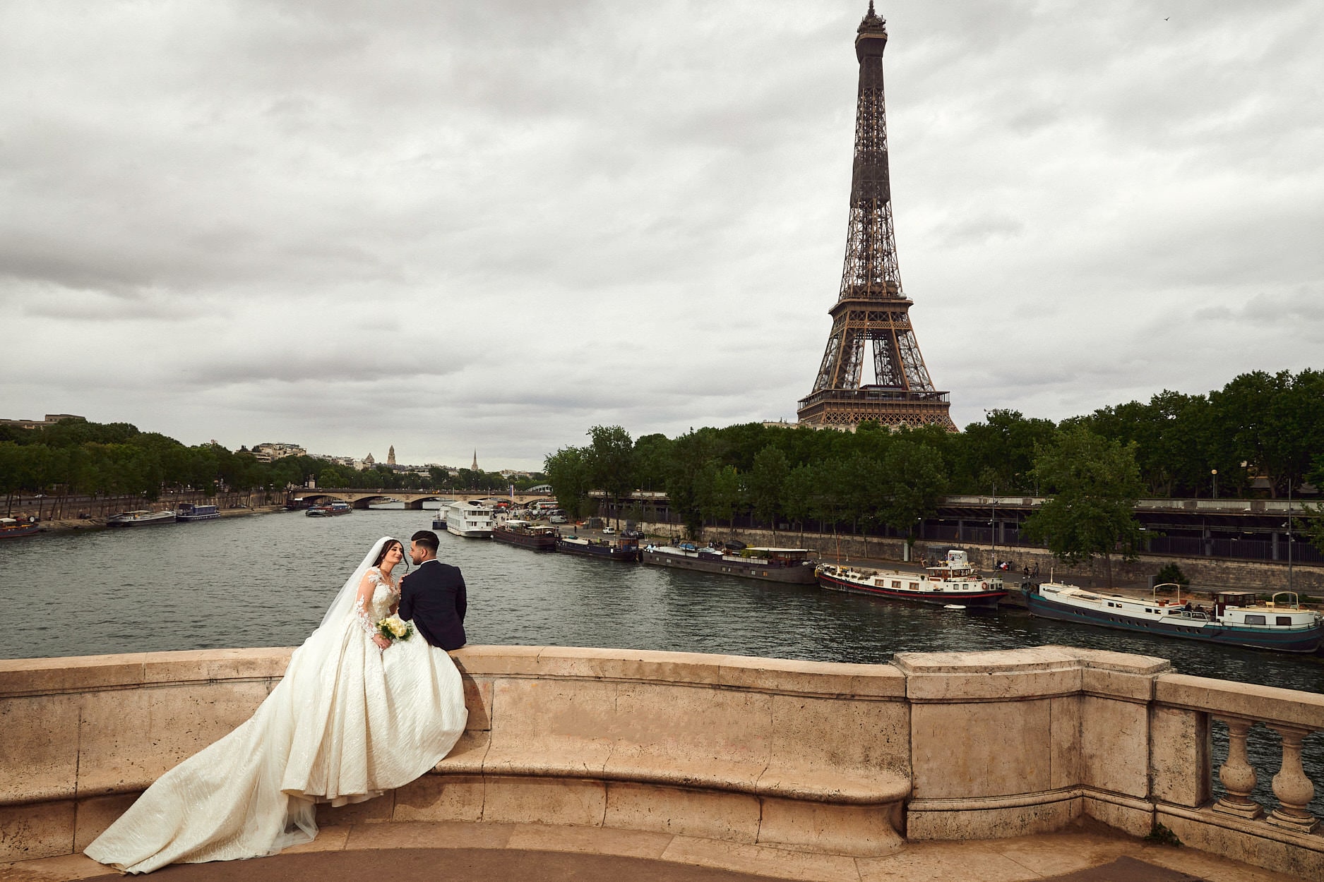 A bride and groom sitting on the curved stone balustrade of a bridge above the Seine, her train spread down the steps, barges moored along the river and the Eiffel Tower behind under a grey sky