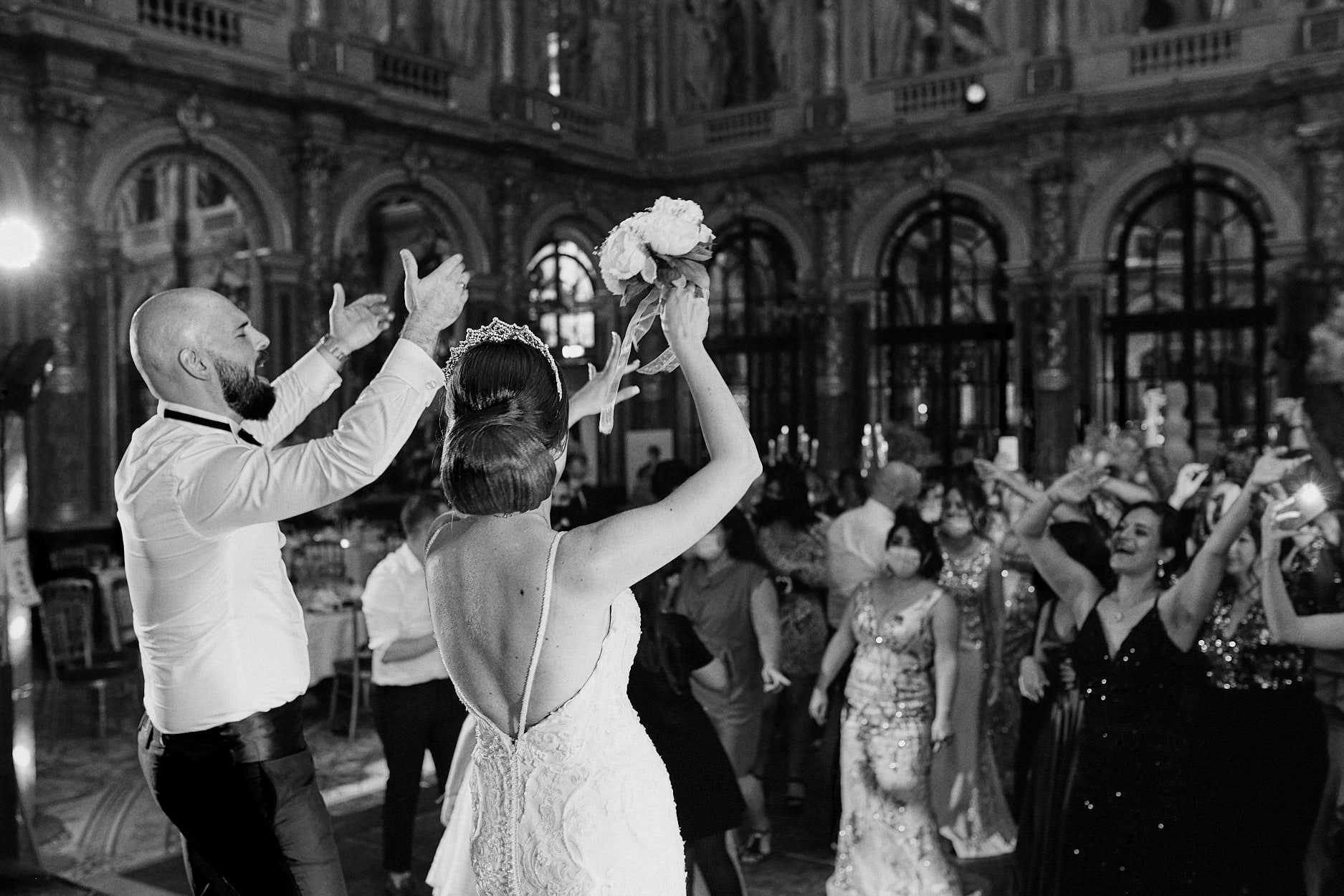 Guests reaching upward as the bouquet leaves the bride’s hand in a grand hall