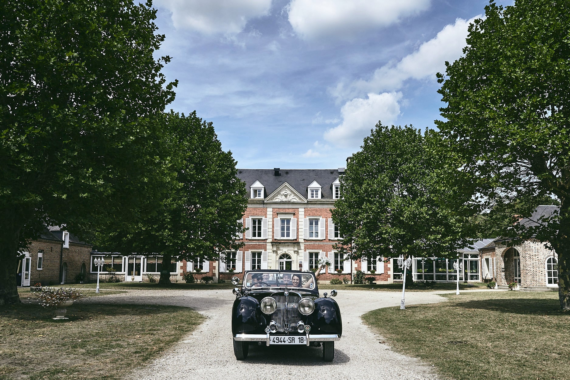 A vintage open-top car waiting on the gravel drive of a château, an avenue of plane trees on either side