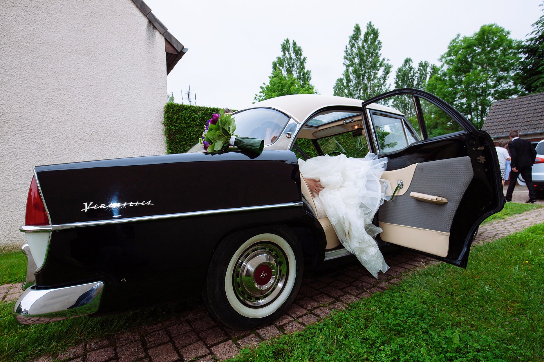 A bride gathering her dress into the back of a black and cream vintage car, the door held open and her bouquet resting on the boot