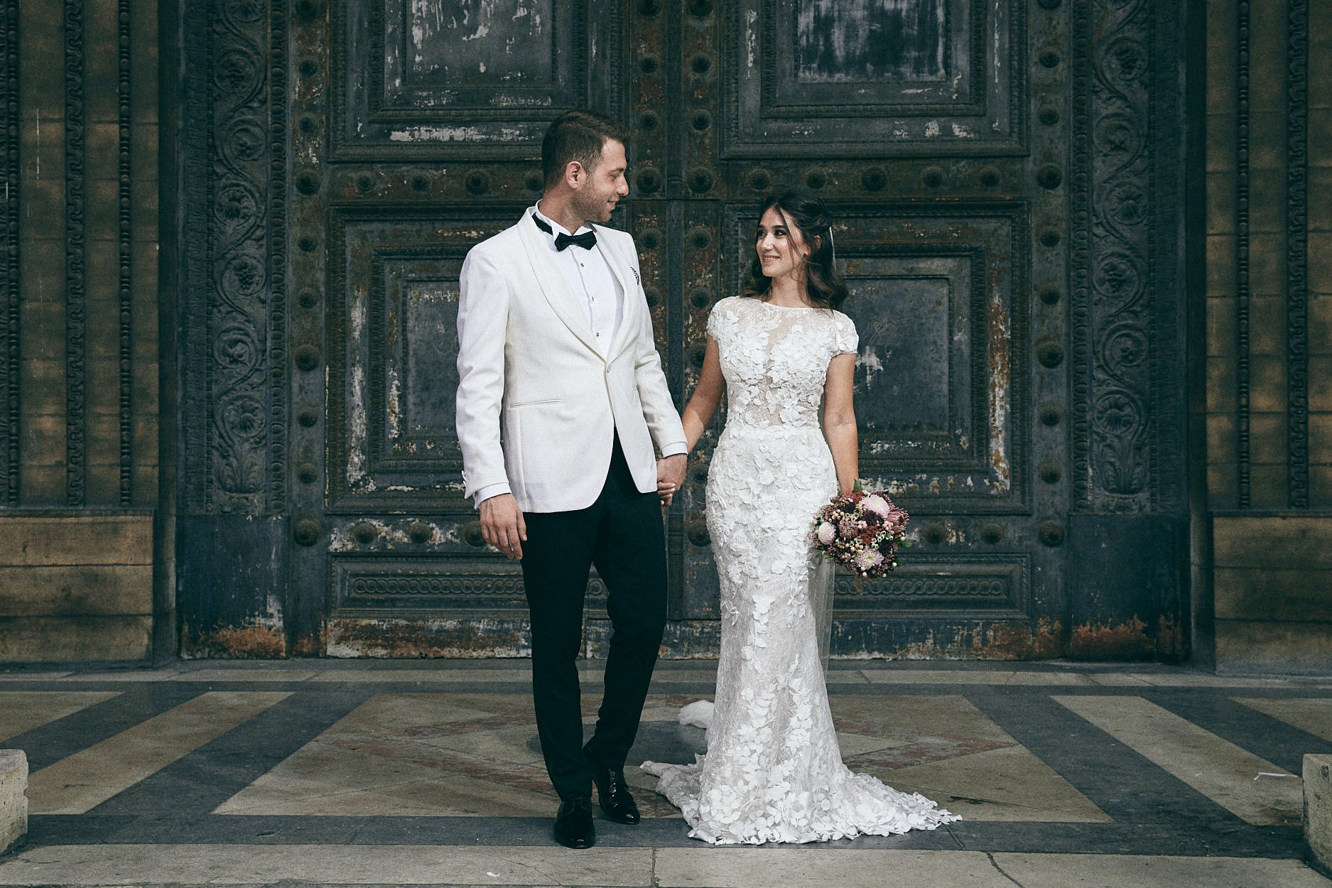 A couple standing before a pair of tall carved bronze church doors, the groom in a white dinner jacket