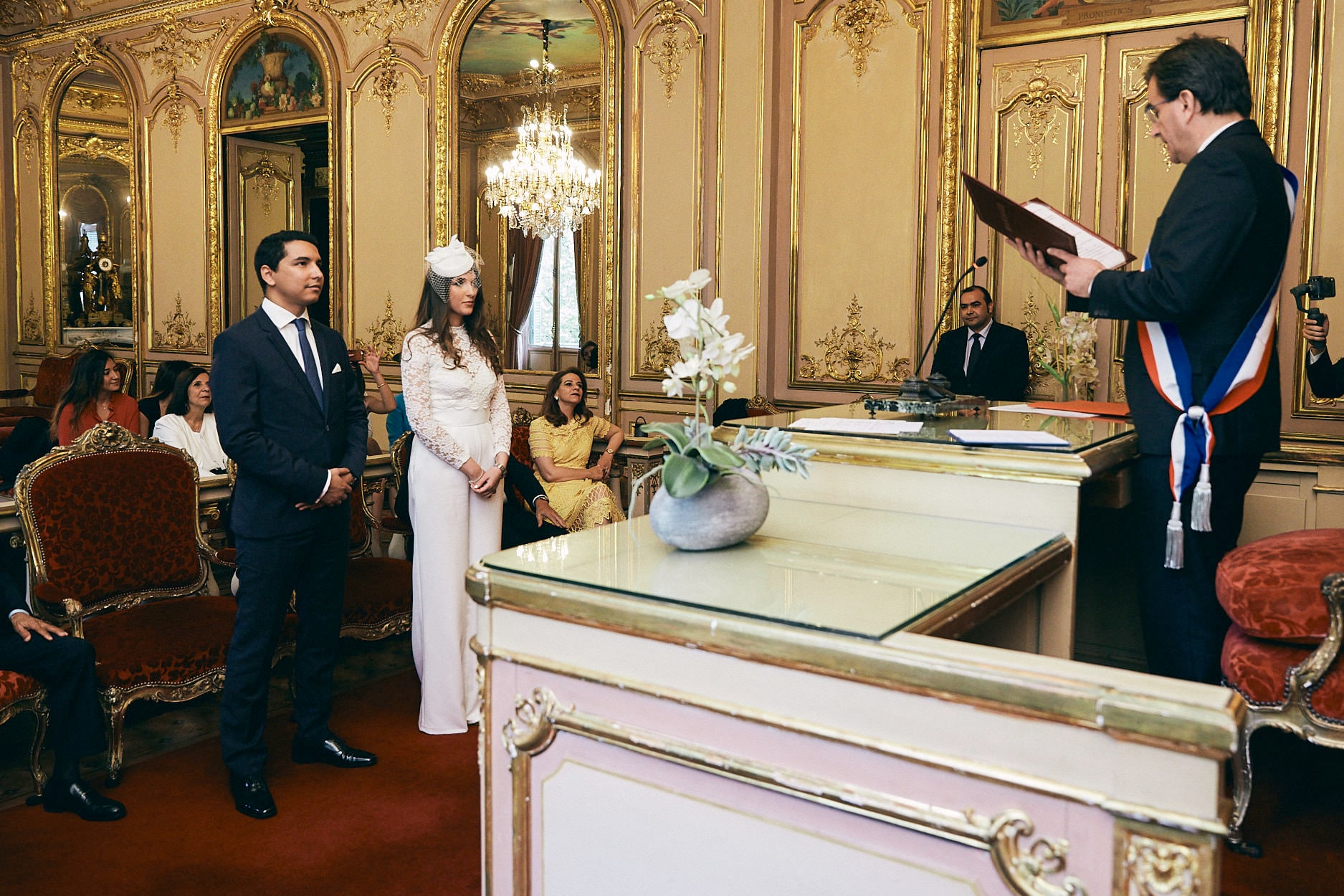 A registrar in his tricolour sash reading to a couple across the desk of a Paris mairie