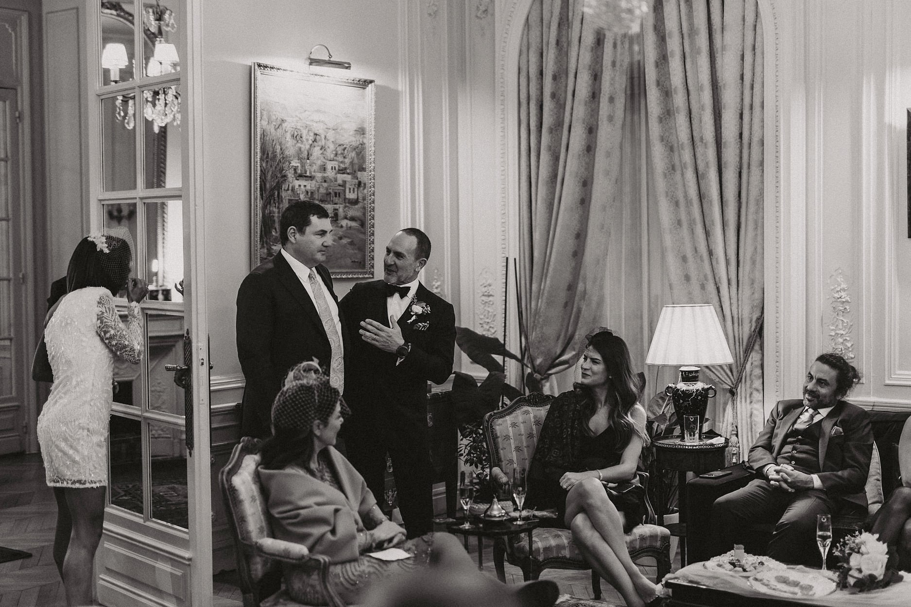 The groom talking with a guest in a panelled drawing room in black and white, others seated around them with champagne, a woman at a mirror in the doorway