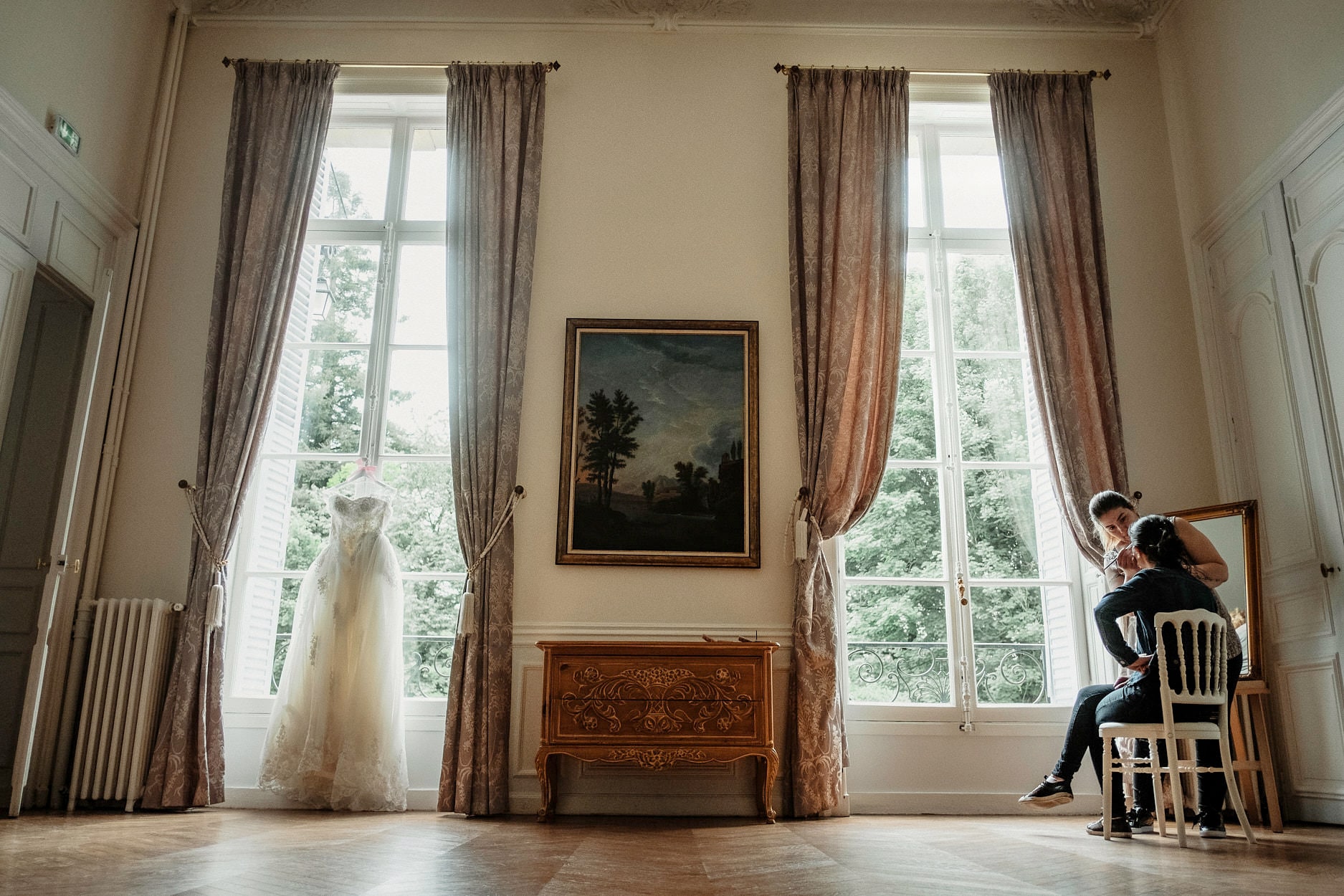 A wedding dress hanging in the window of a tall panelled room, the couple sitting together in the far corner