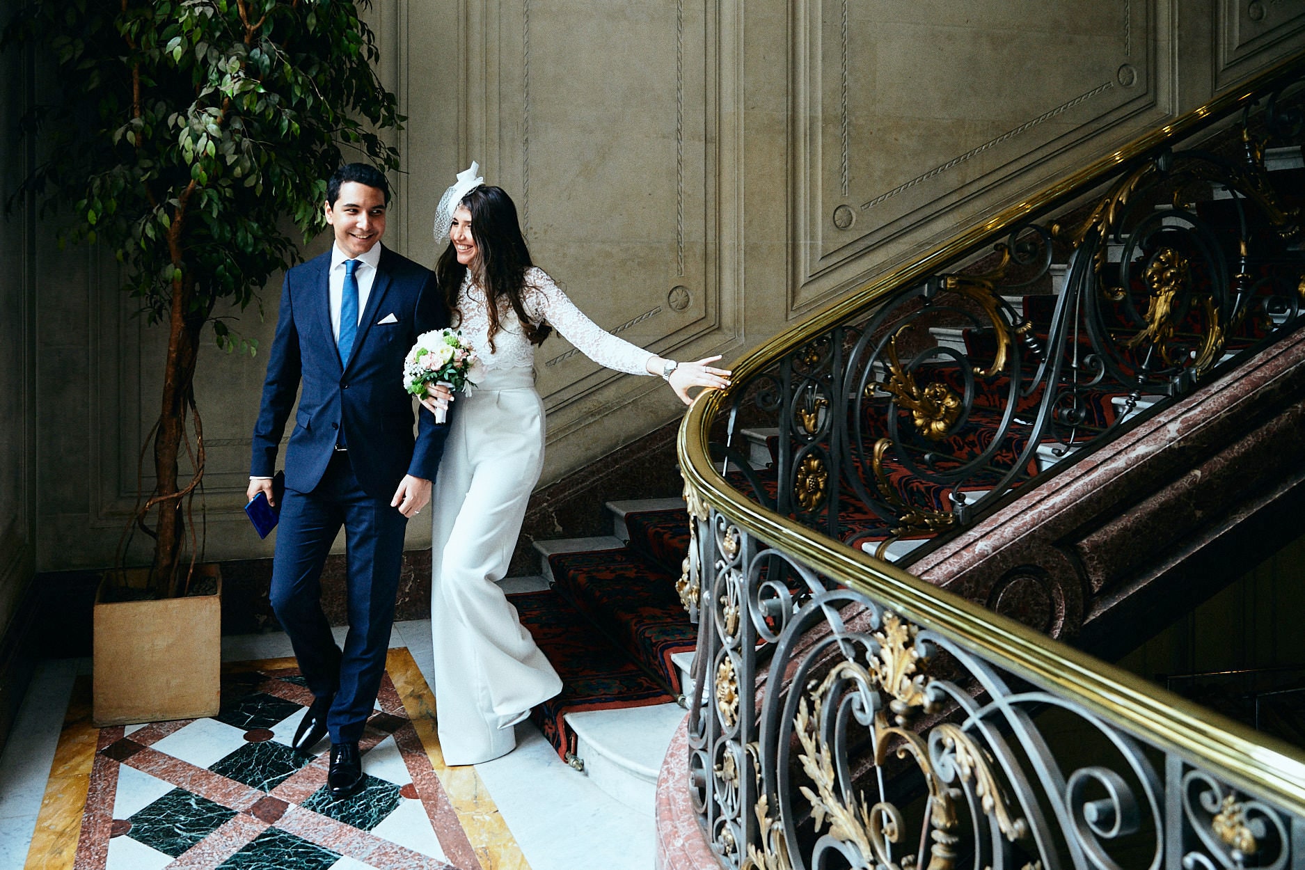 A bride and groom coming down a gilded town hall staircase, her hand on the brass rail, his blue suit against the panelling