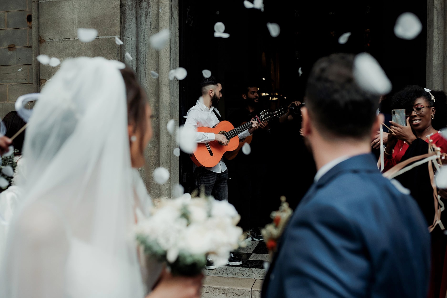 A guitarist playing in a dark doorway as confetti falls, the bride and groom watching from the front of the frame