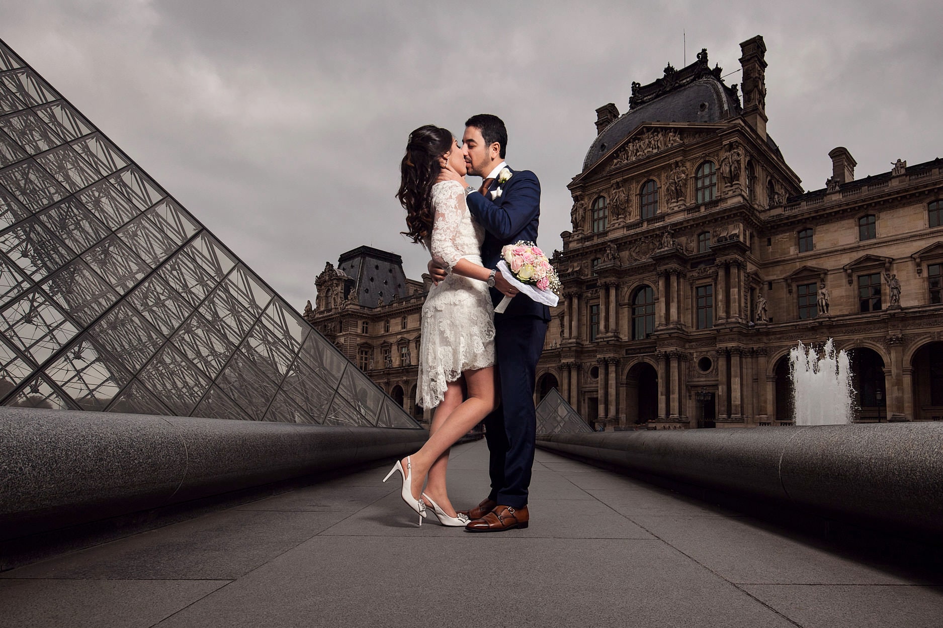 A bride and groom kissing in the courtyard of the Louvre, the glass pyramid on one side of them and the palace on the other, under a heavy grey sky