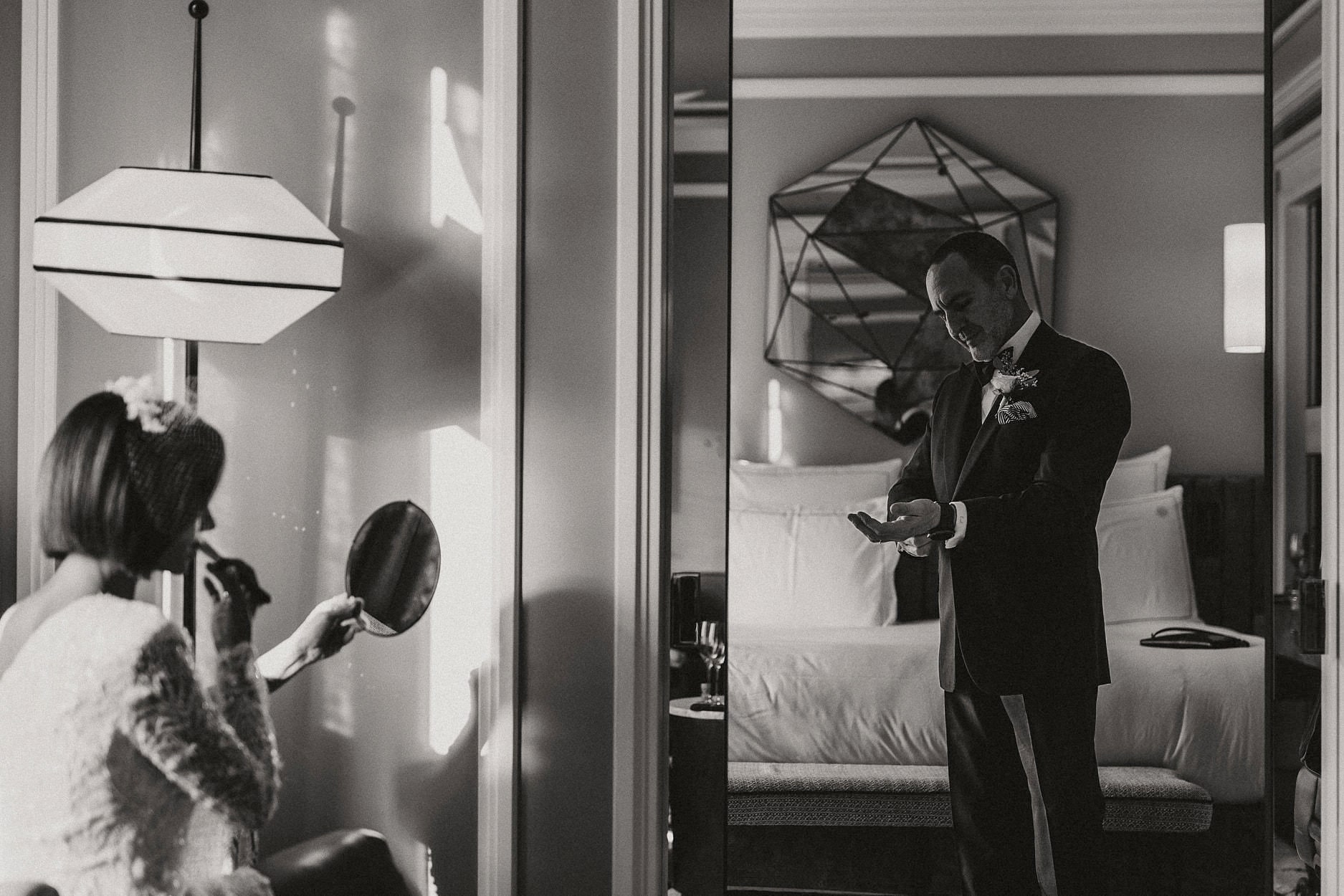 A groom seen through a doorway in a hotel room while a bridesmaid checks her reflection
