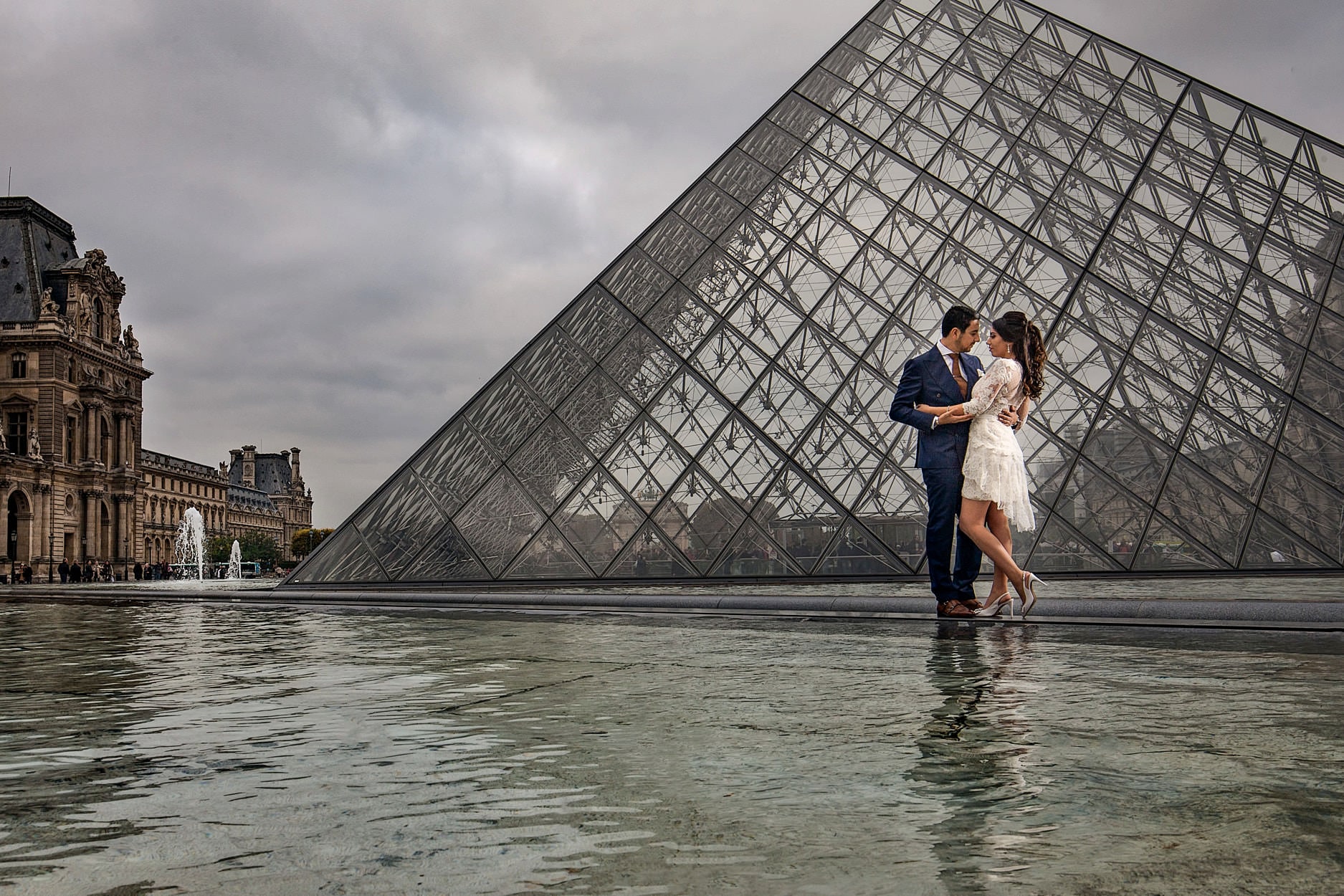 A bride and groom standing in the shallow pool in front of the Louvre pyramid, reflected in the water, under an overcast sky
