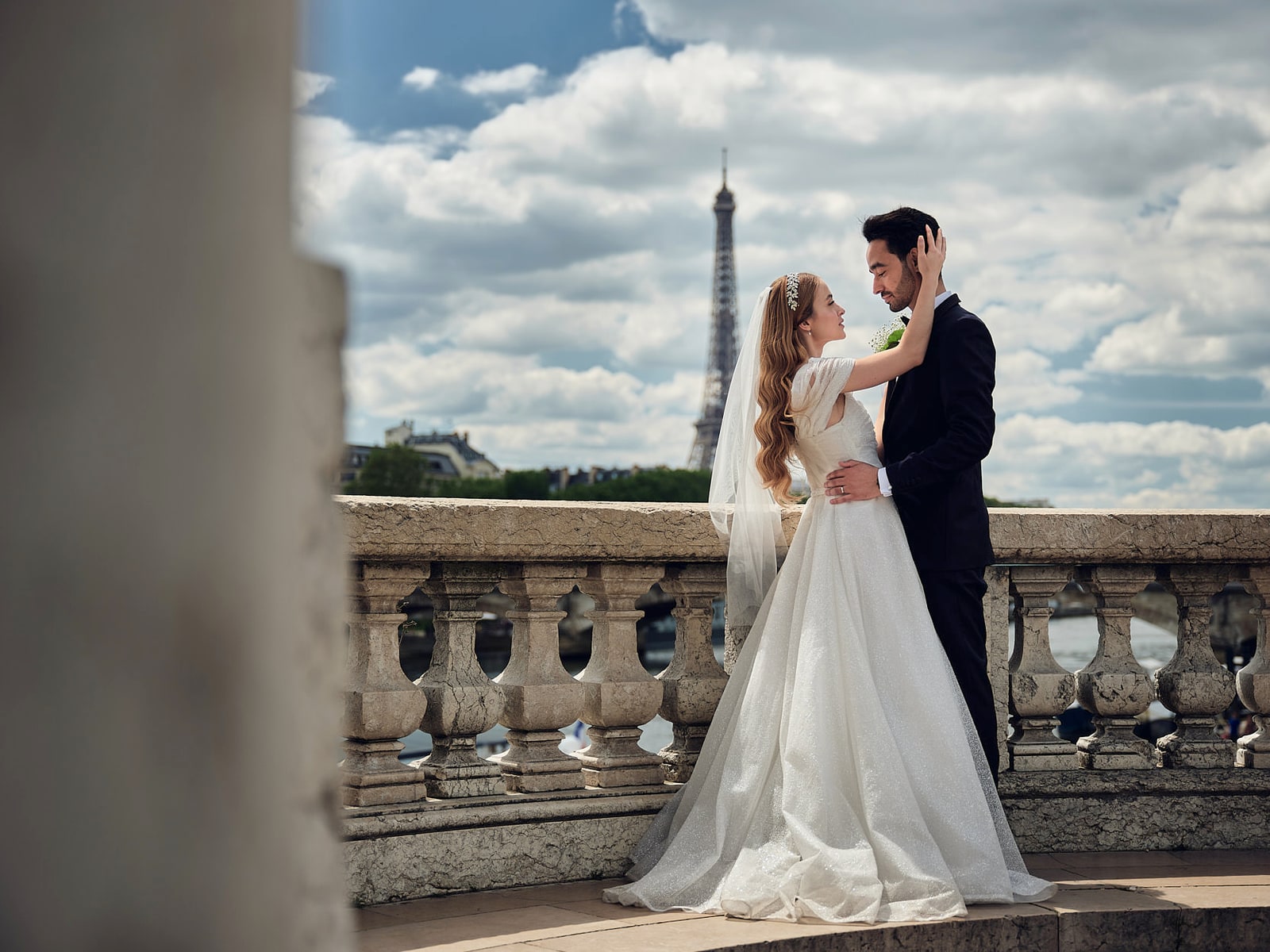 A bride and groom leaning on a stone rail above the river with the Eiffel Tower behind them