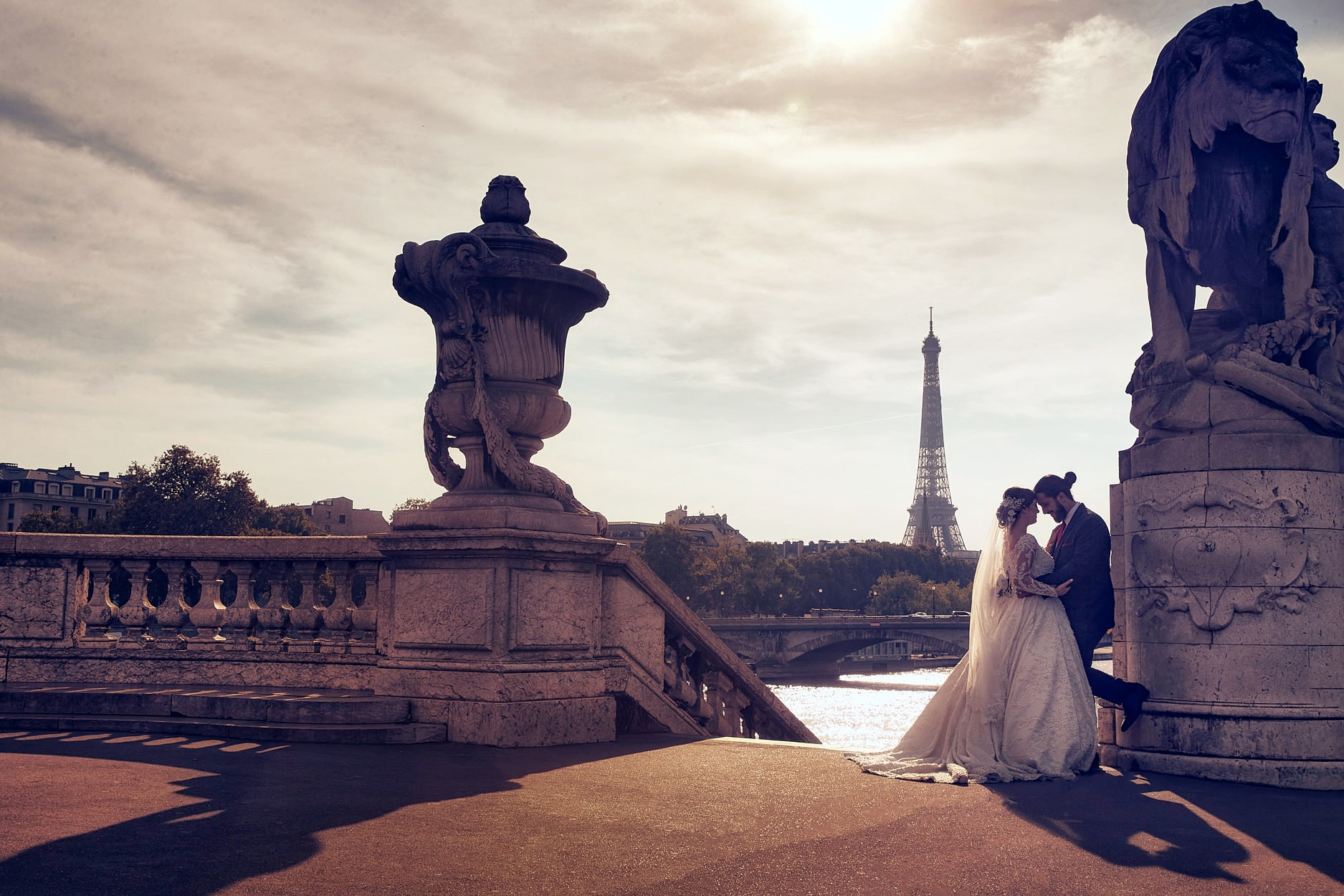 A bride and groom forehead to forehead on the Pont Alexandre III at golden hour, a carved stone urn on one side and a lion on the other, the Eiffel Tower across the Seine between them