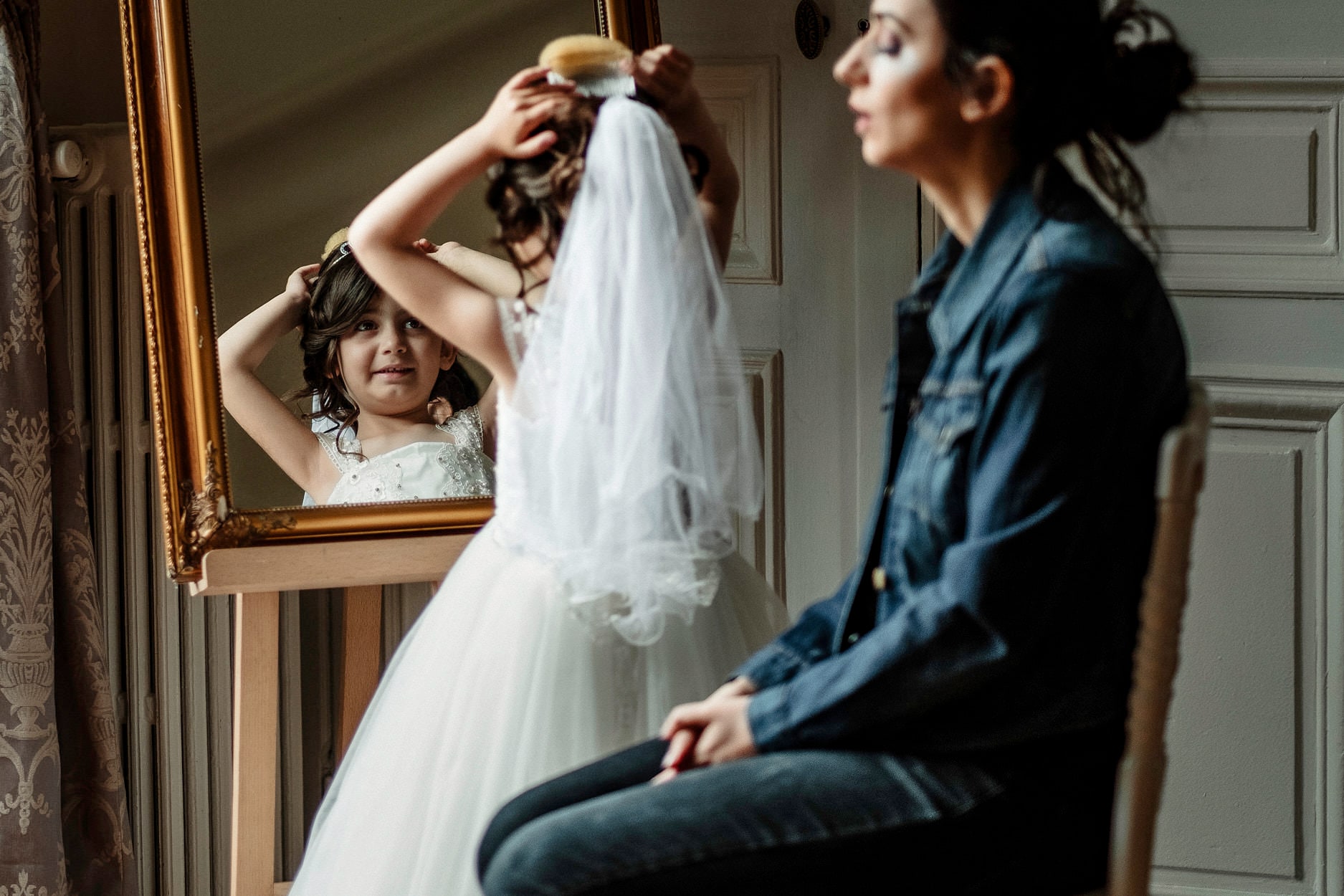 A woman lifting a veil towards a bride reflected in a gilt mirror