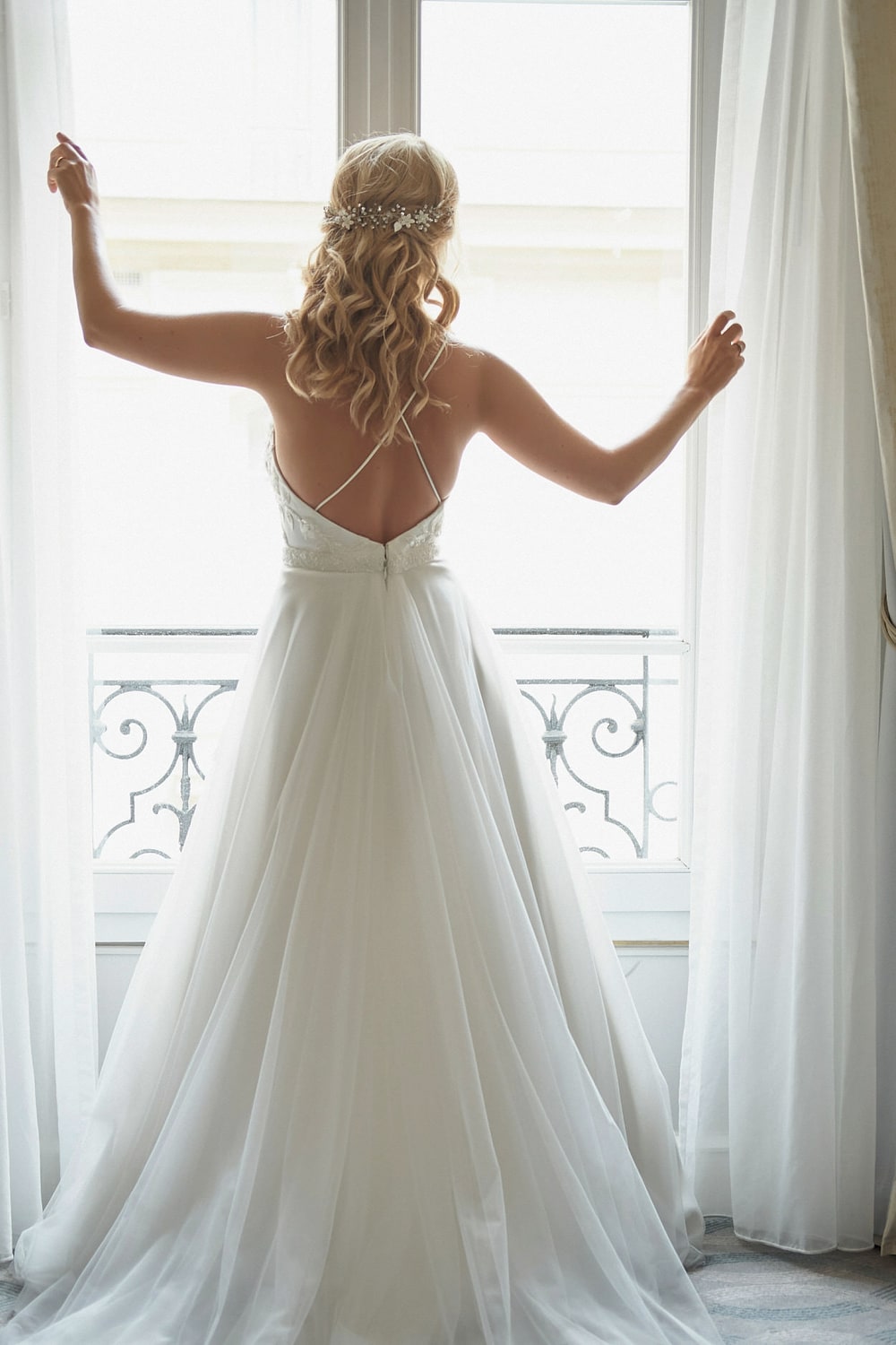 A bride standing at a tall window with both hands on the curtains, seen from behind against the light