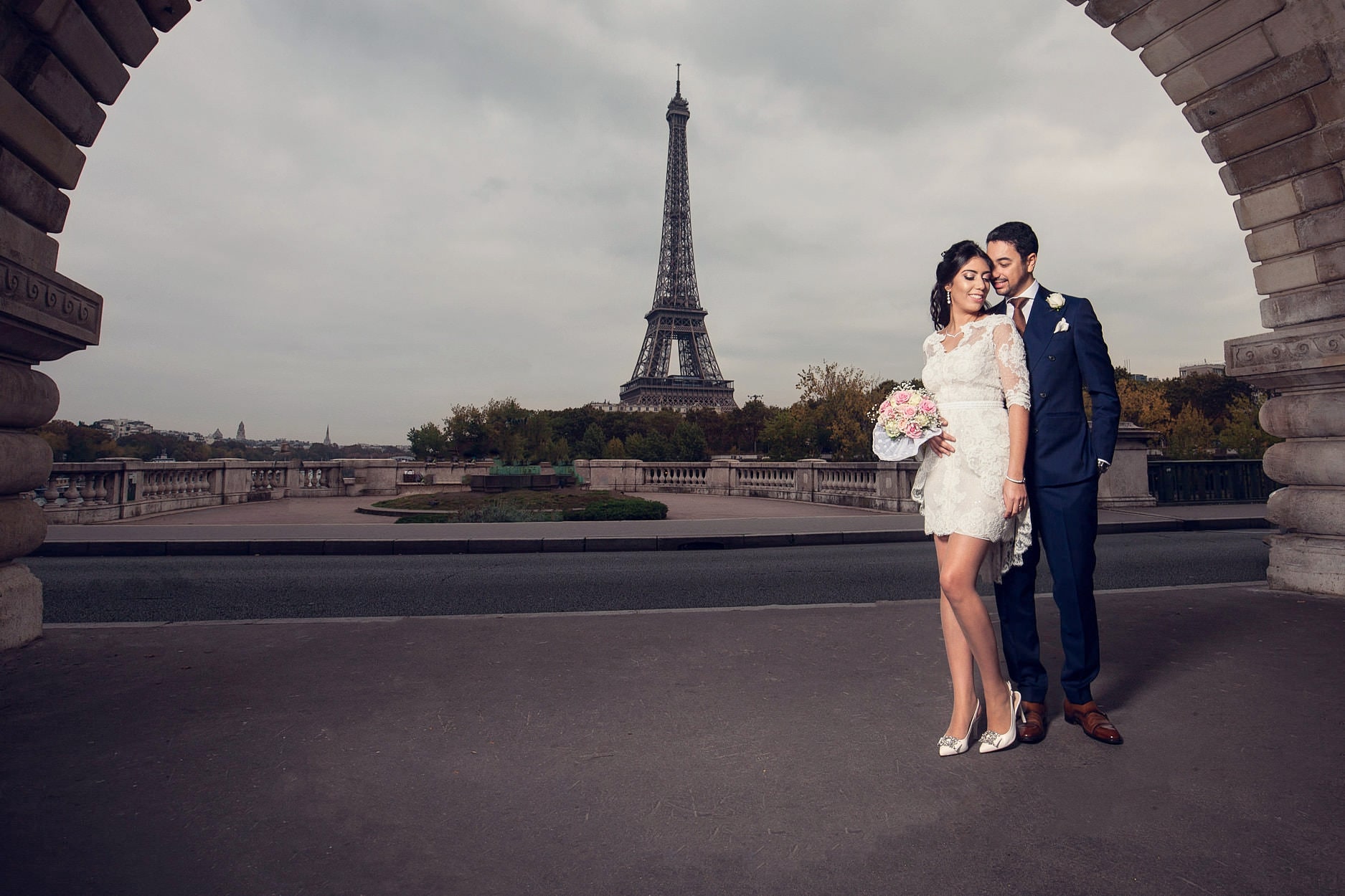 A couple standing under a stone arch of the Pont de Bir-Hakeim with the Eiffel Tower framed beside them