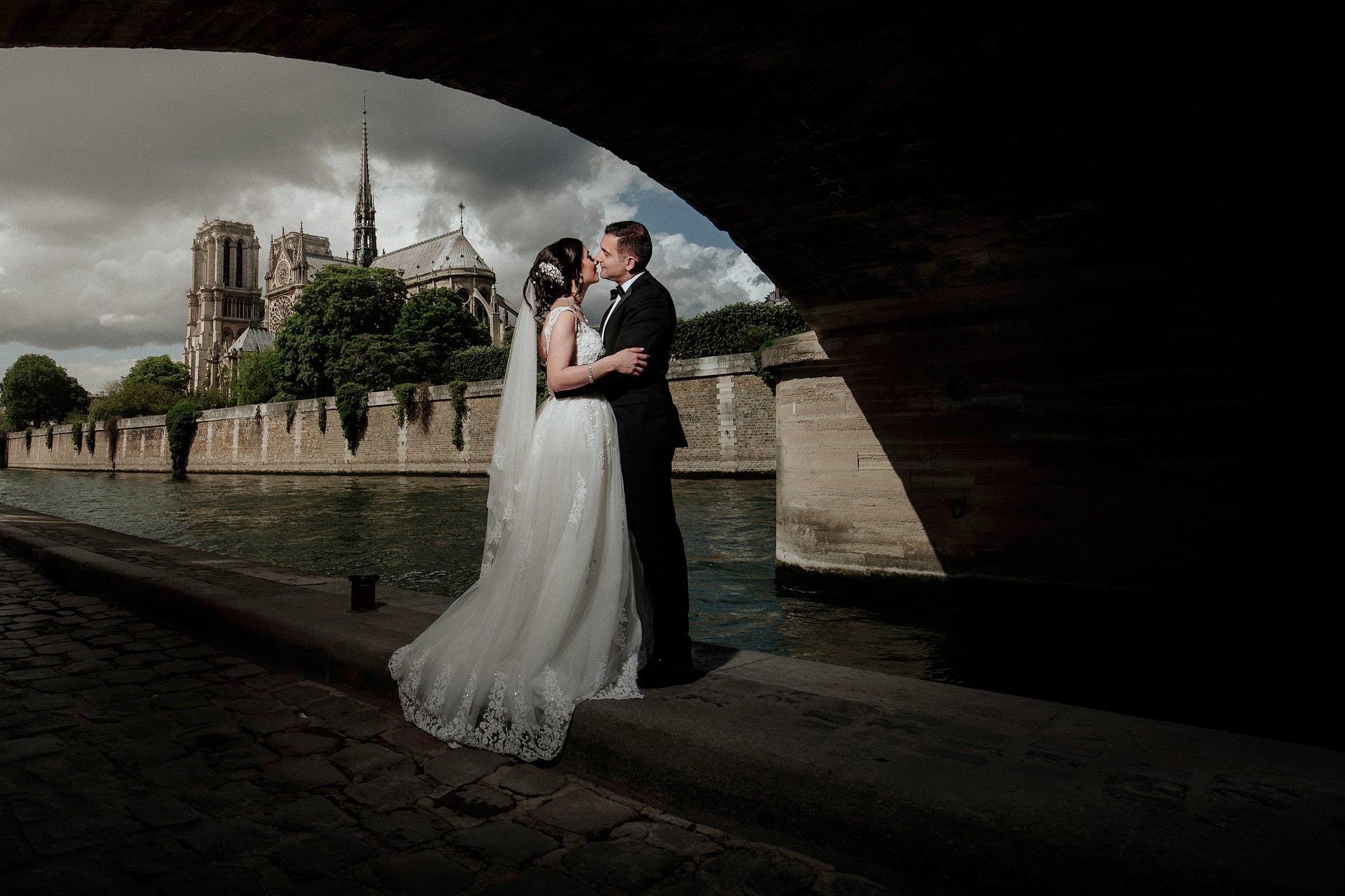 A bride and groom kissing on the quay beneath a bridge arch, Notre-Dame and the Seine beyond them