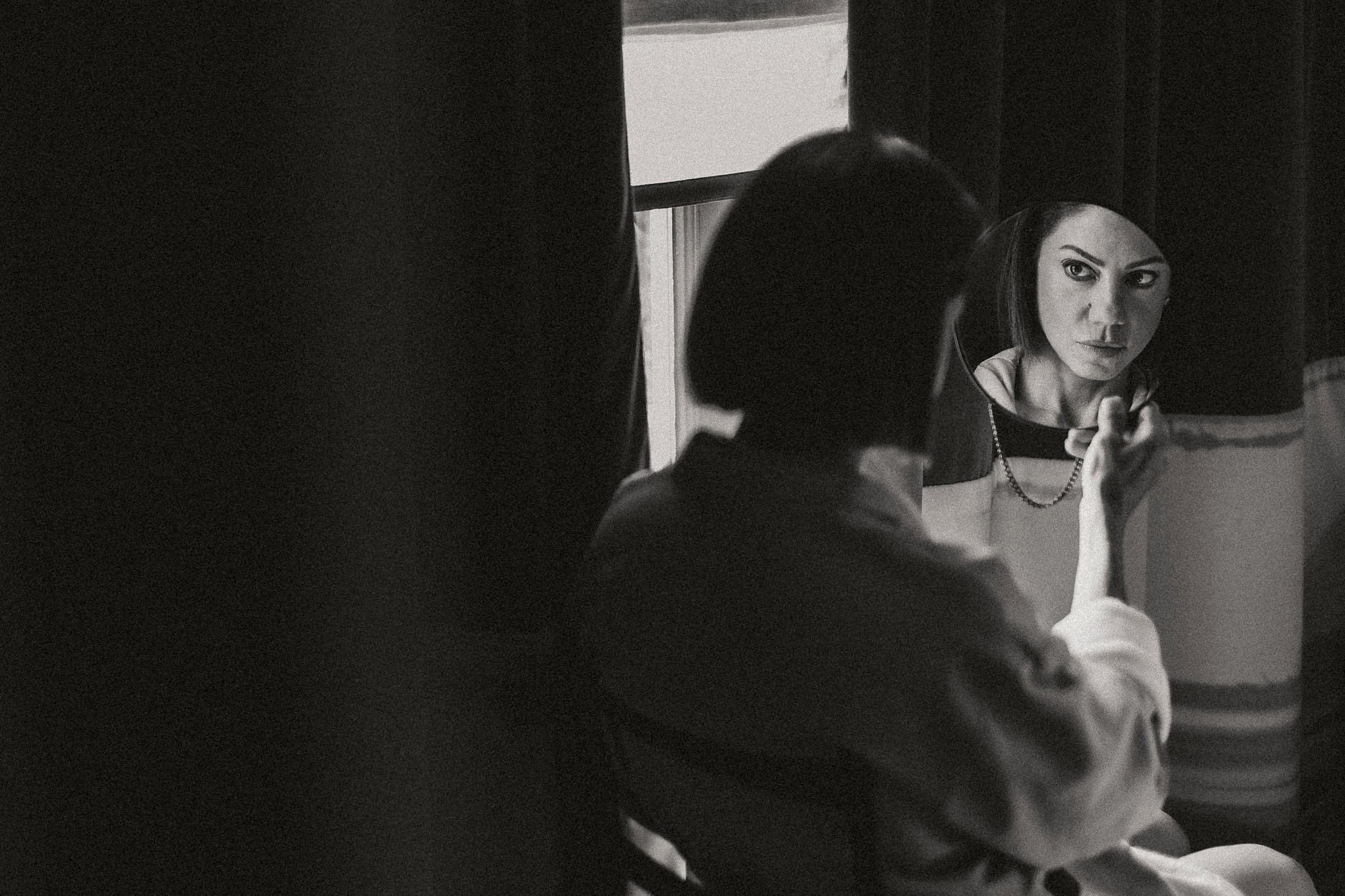 Two women talking quietly by a hotel window in low light, one still in a dressing gown